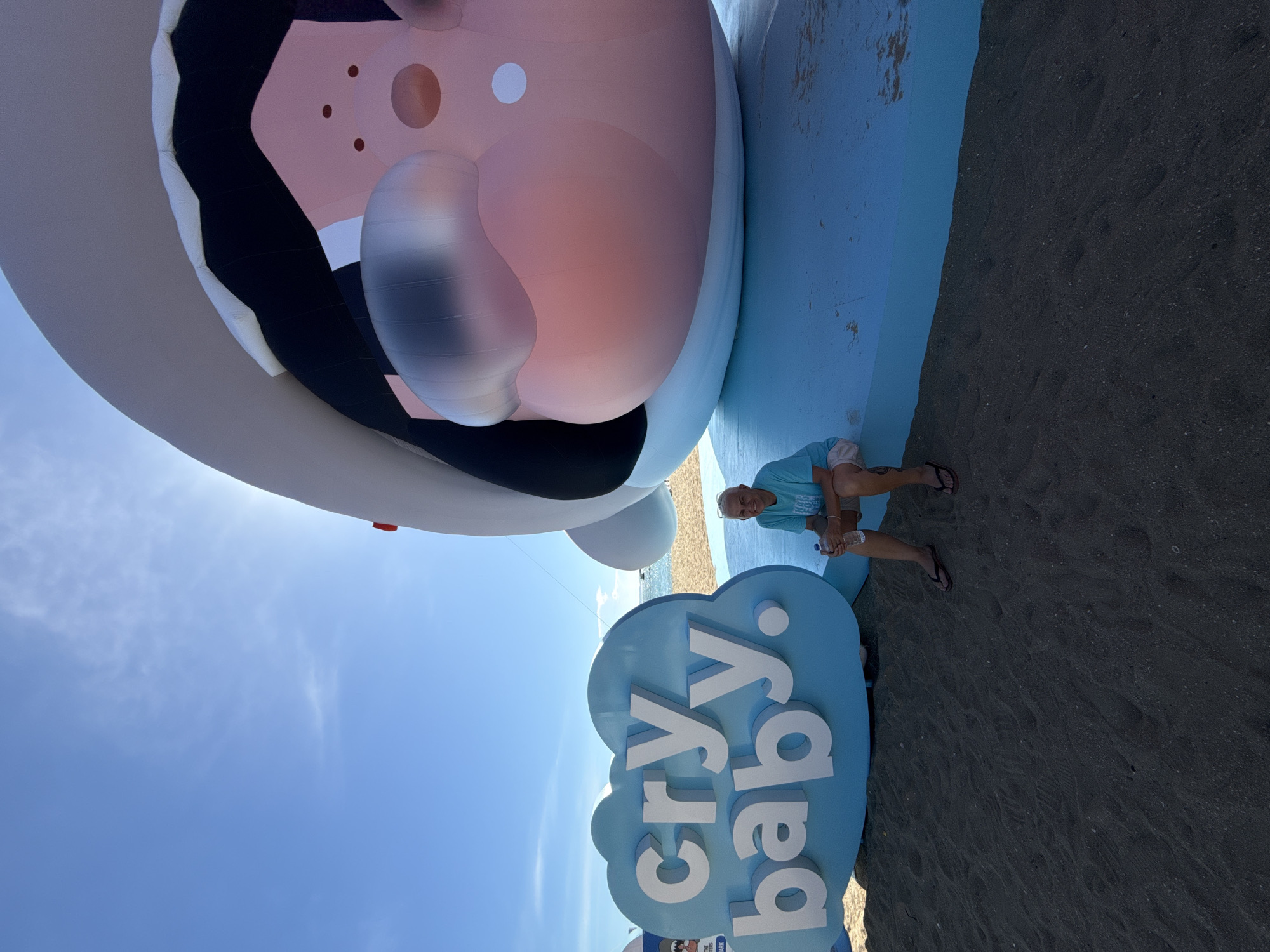 A person is crouching on the beach next to a large, inflatable cartoon character and a blue cloud-shaped sign. The character is white and pink with black and brown accents, and appears to be a baby with a large head. The sign reads "cry baby." The person is wearing a blue t-shirt and shorts and is holding a bottle of water. The scene is outdoors on a sunny day, with the ocean visible in the background. The sand appears to be dark, and there are some footprints visible. The sky is blue with a few wispy clouds. The overall mood appears to be relaxed and playful. The location is Pattaya City, Thailand.
