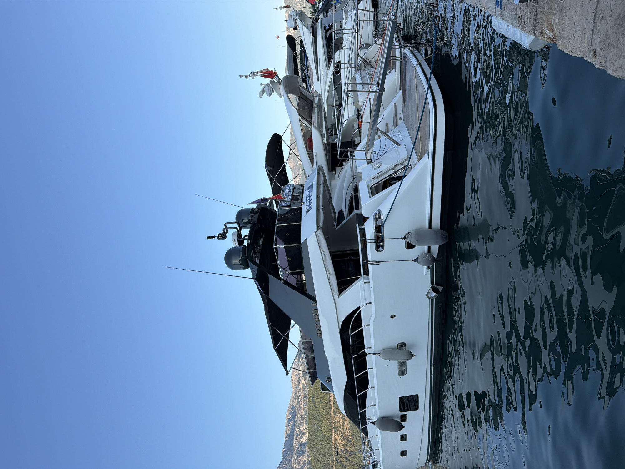 In the city of Budva, Montenegro, a fleet of luxury yachts are moored in a marina under a clear blue sky. The image focuses on a large white yacht in the foreground, which has a sleek, modern design with black accents on its upper deck and canopy. The yacht features multiple levels, windows, and railings, and is equipped with gray fenders along its side. Behind it, several other yachts of similar style are docked, creating a line of impressive vessels. The water in the marina is dark and reflects the shapes of the boats and the sky. In the background, rolling hills covered with greenery rise up, with a clear horizon line against the bright sky. The overall scene depicts a tranquil and opulent setting within the coastal city.