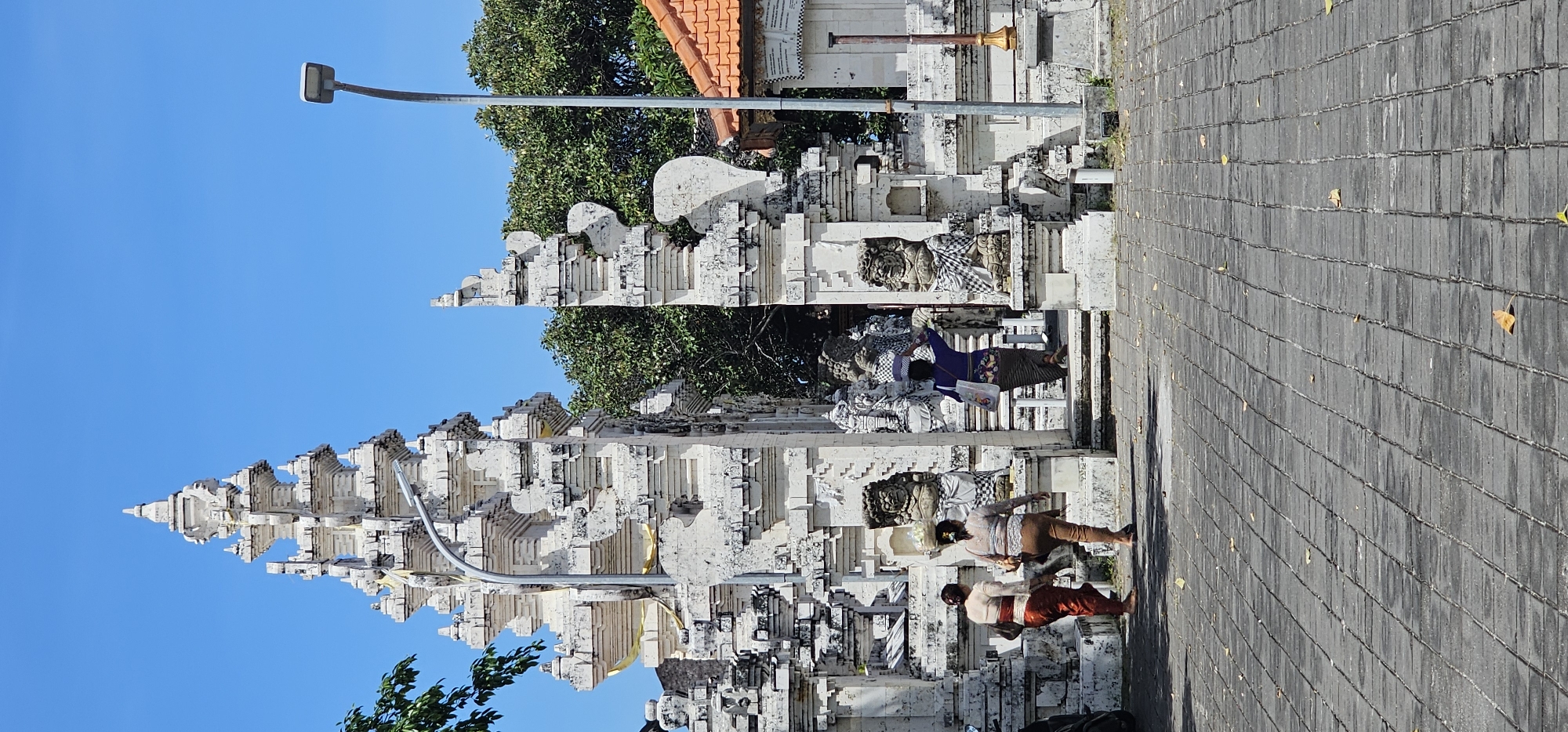 The image shows a traditional Balinese gate or temple entrance in Kuta Selatan, Indonesia. The structure is white and layered, with intricate carvings. A lamp post stands nearby. Several people are walking in front of the gate. The ground is paved with gray stones. The sky is blue. Trees are visible behind the gate.