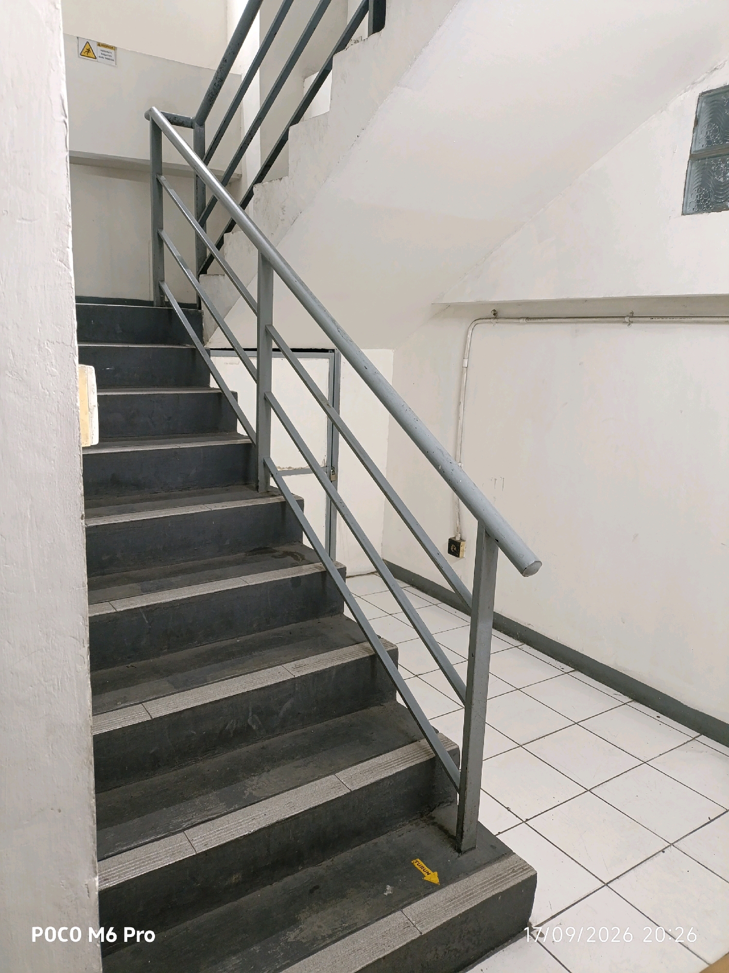 The image depicts a concrete staircase with a metal railing. The stairs are dark gray with lighter gray textured strips on the edge of each step. The walls are painted white. There is a yellow directional arrow on the bottom step pointing downwards. The floor is tiled with white square tiles. A small, frosted glass window is visible in the upper right corner of the image. Electrical conduits run along the wall near the ceiling. A warning sign with a yellow triangle and black exclamation mark is affixed to the upper left wall. The timestamp "17/09/2026 20:26" and the text "POCO M6 Pro" are present in the lower right and lower left corners of the image, respectively.