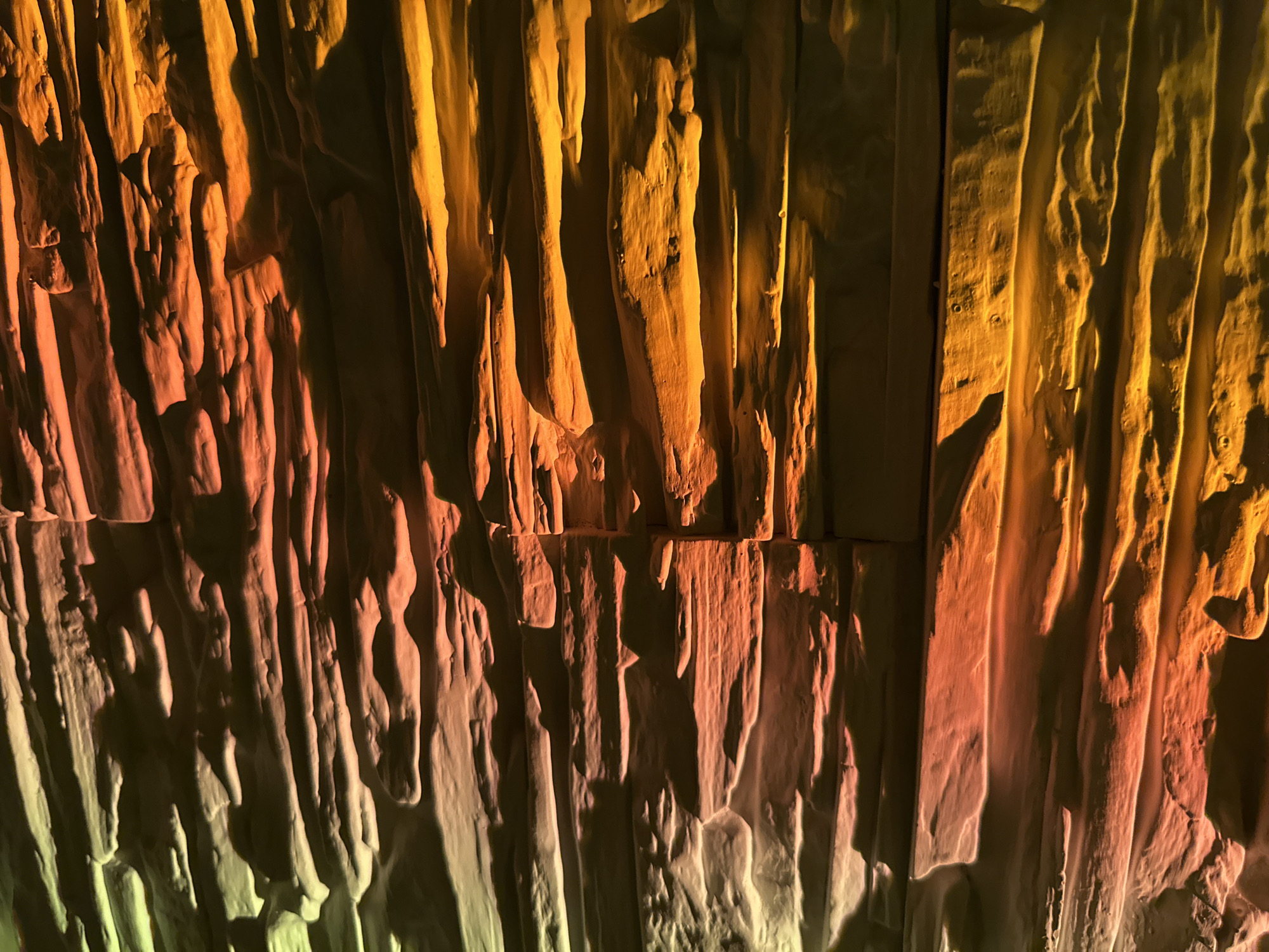 This image presents a detailed view of a highly textured surface, dramatically illuminated by artificial colored lights. The surface features pronounced vertical ridges, grooves, and undulating forms that create a rough, uneven relief with deep, sharp shadows. These shapes vary in width and depth, strongly resembling natural rock formations such as stalactites, flowstone, or columnar basalt. Alternatively, it could be an architectural or artistic installation designed to mimic such organic textures, possibly crafted from carved plaster, wood, or a similar material. A distinct horizontal line, potentially indicating a seam between panels or a natural break in the formation, is visible across the mid-right section of the frame.
The lighting source casts a vibrant color gradient across the textured surface. The upper and left portions of the image are bathed in warm reddish-orange and amber tones, which seamlessly transition to cooler, yellowish-green hues towards the bottom and right. This multi-colored illumination accentuates the three-dimensional contours and deep shadows, enhancing the visual depth and tactile quality of the texture.
No people, animals, or specific objects are present in the image, and no activity or event is taking place. There is no visible text. The sophisticated and controlled nature of the lighting suggests an indoor setting, such as an art gallery, a themed exhibition space, or a decorative architectural feature within a commercial or public building. The time of day is not indicated, but the artificial illumination implies an indoor environment. The overall impression is one of abstract beauty, highlighting the interplay of texture, light, and color.