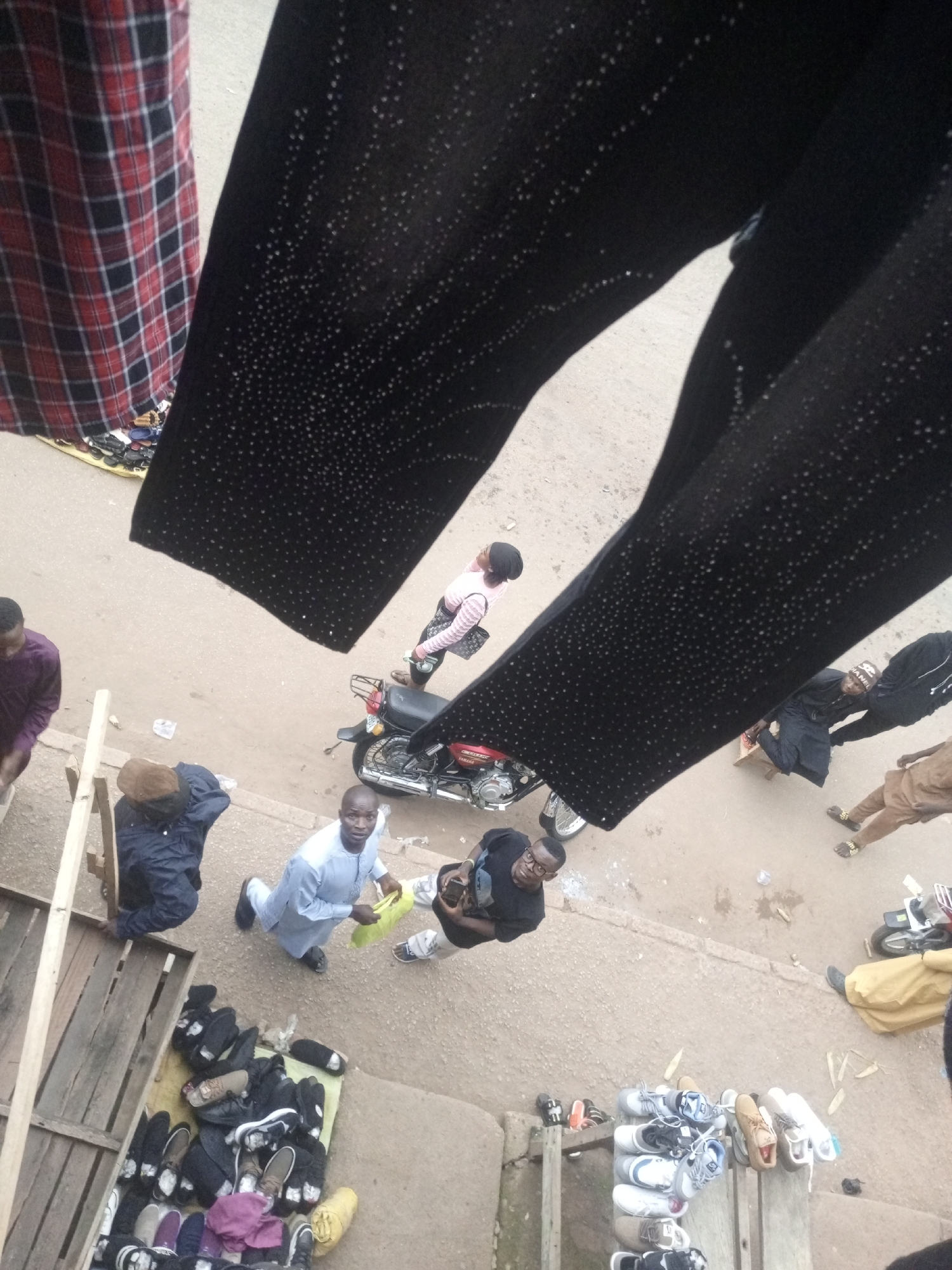 This is an overhead shot of a bustling street market in Bukuru, Nigeria. The foreground is dominated by a person's black pants adorned with a scattering of tiny, reflective dots. To the left, a red and black plaid fabric is visible, likely clothing or a textile display.
The scene below is a snapshot of daily life. Several individuals are present. On the left, a man in a purple garment stands near a wooden structure, possibly a market stall. Beside him, another person in a dark blue jacket and brown hat is seated. In the center, a man in a light blue traditional outfit is kneeling, holding a yellow bag, and looking towards the camera. To his right, a man in a black t-shirt and glasses is also looking upwards. Further right, two people in traditional attire, one in brown and another in dark clothing, are walking away. A red motorcycle is parked nearby.
Various goods are displayed for sale. A collection of shoes and sandals is arranged on the ground and on wooden planks. The ground is a dusty, earthy surface, typical of an outdoor market. The overall impression is one of vibrant activity and commerce.