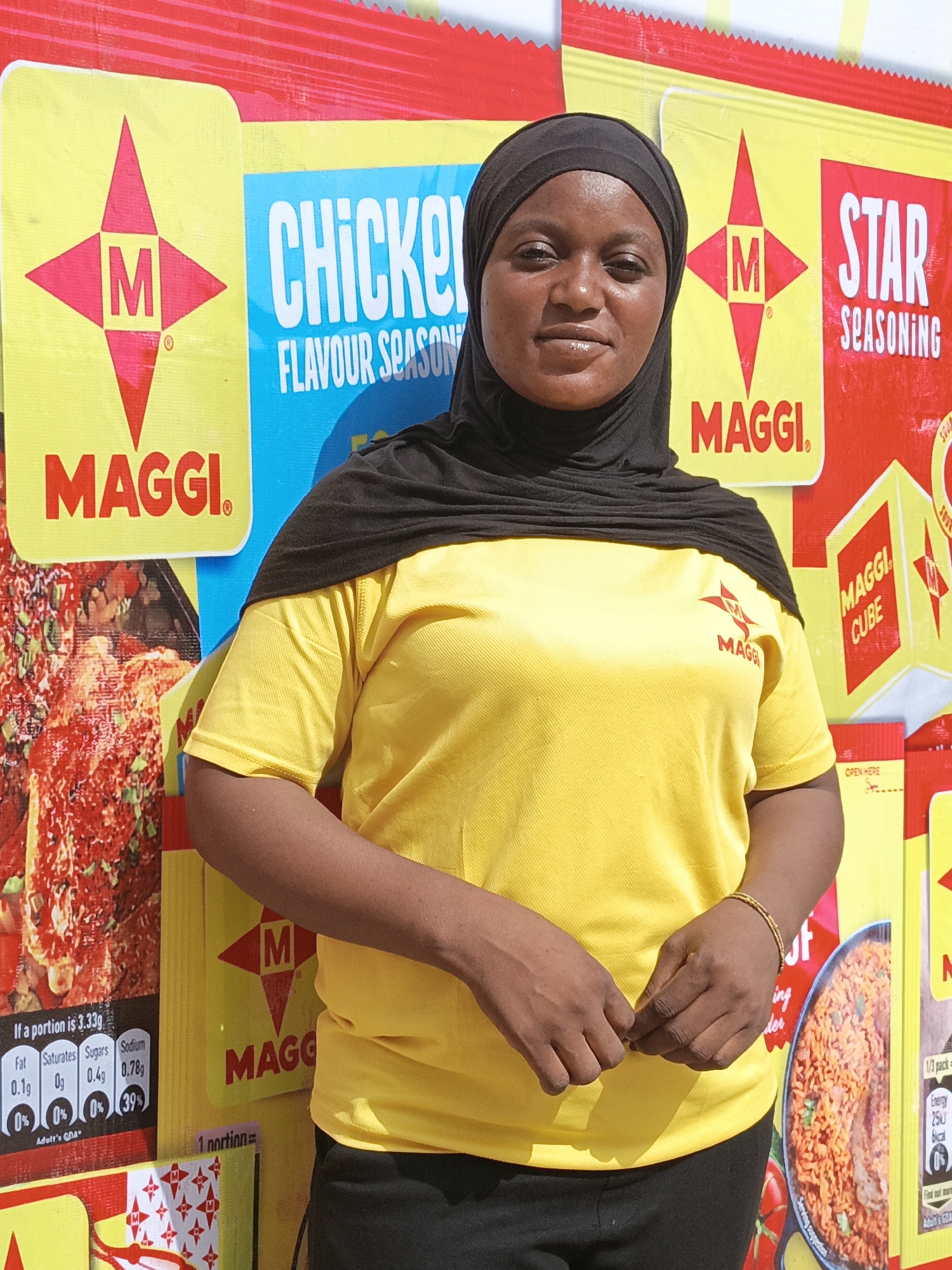 A woman is pictured standing outdoors, likely at a promotional event or a market stall in Kawaji, Nigeria, given the bright daylight and the commercial backdrop. She has dark skin and is wearing a black hijab that covers her head and neck, and a bright yellow short-sleeved t-shirt. The yellow t-shirt features a red Maggi "M" star logo on the left chest. Her hands are clasped in front of her, and she wears a thin gold-colored bracelet on her right wrist. She looks directly at the camera with a neutral expression.
The background is dominated by a large, colorful display featuring Maggi brand products. The display mimics large packets of Maggi seasoning, primarily in yellow, red, and blue colors. Prominently visible are the "MAGGI" brand name and its distinctive "M" star logo. Text on the display includes "CHICKEN FLAVOUR SEASONING" on a blue panel, and "STAR SEASONING" on a red panel. Another section shows "MAGGI CUBE."
Detailed nutritional information is visible on a dark panel, stating: "If a portion is 3.33g," followed by values for "Fat 0.1g 0%," "Saturates 0g 0%," "Sugars 0.4g 0%," and "Sodium 0.78g 39%," with "Adult's GDA" indicated. Other partial text includes "1 portion=", "OPEN HERE," and "Energy 25kJ 6kcal." Images of food, such as what appears to be jollof rice and a chicken dish, are also part of the vibrant product packaging illustrations on the display.