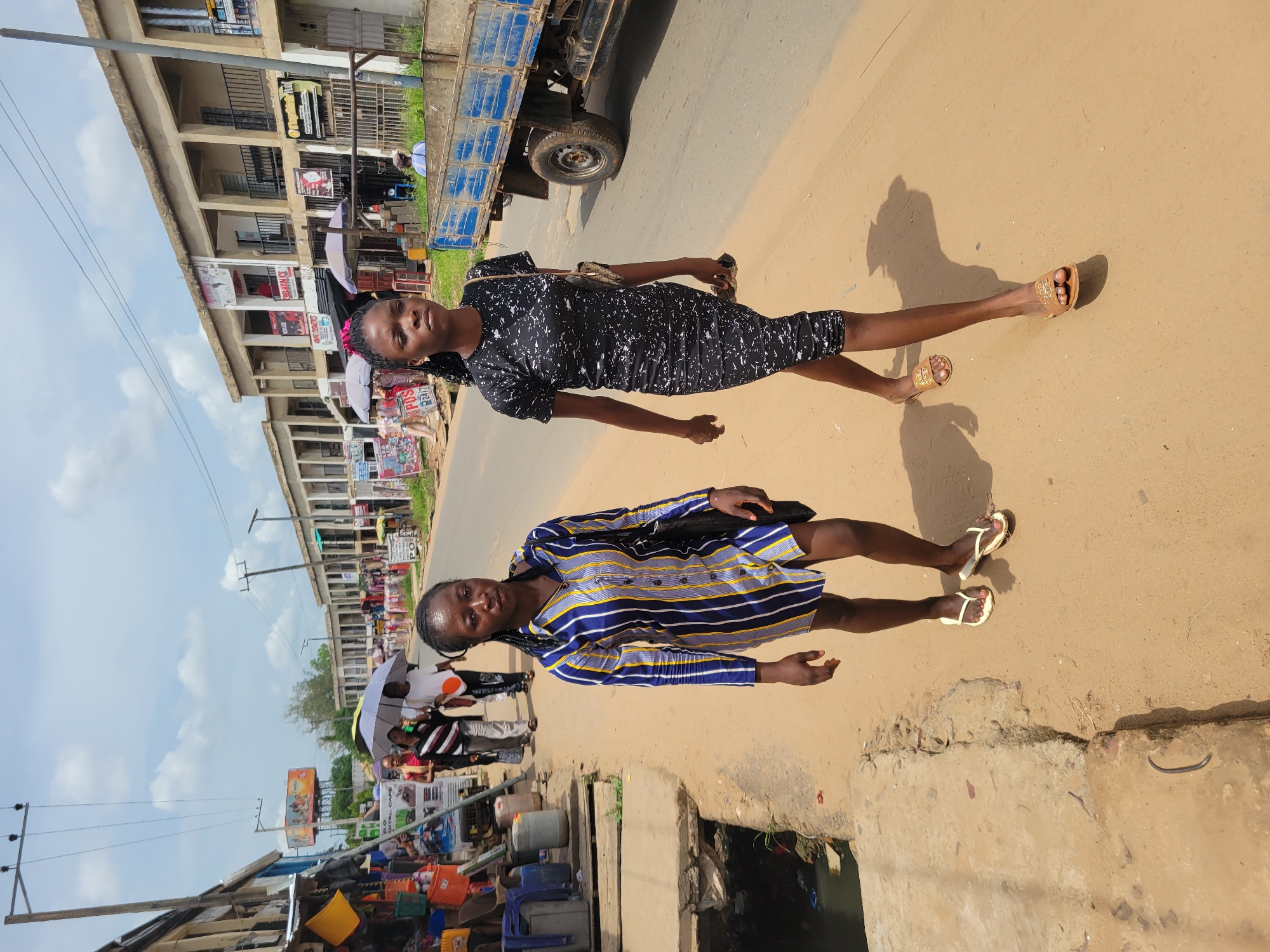 Two young women stand in the street of Egbagu, Nigeria. The woman in the foreground wears a blue and yellow striped dress and white sandals. The woman behind her is wearing a black and white patterned dress and gold sandals. A blue truck is parked on the street nearby. Buildings are visible in the background. A drainage ditch is adjacent to the road. People and vendors are present on the side of the road.