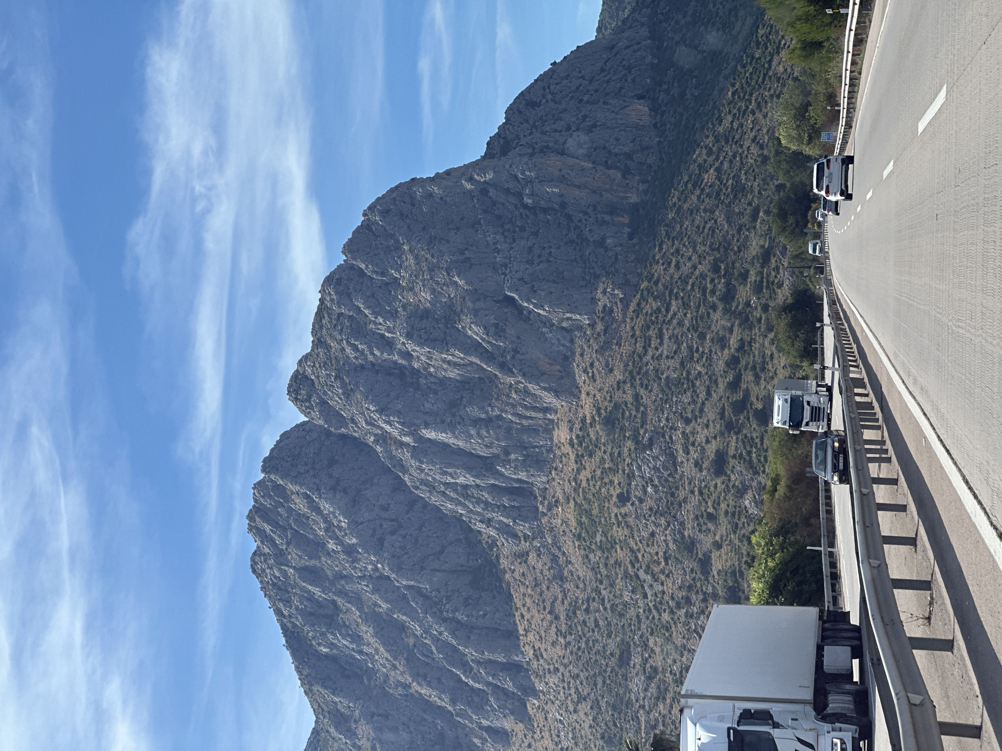 The image depicts a highway scene in Ondara, Spain, with a large mountain dominating the left side. The sky is blue with scattered clouds. A multi-lane highway runs on the right side, bordered by a metal guardrail. Several vehicles, including trucks and cars, are visible on the road. The mountain is rocky and partially covered with vegetation.