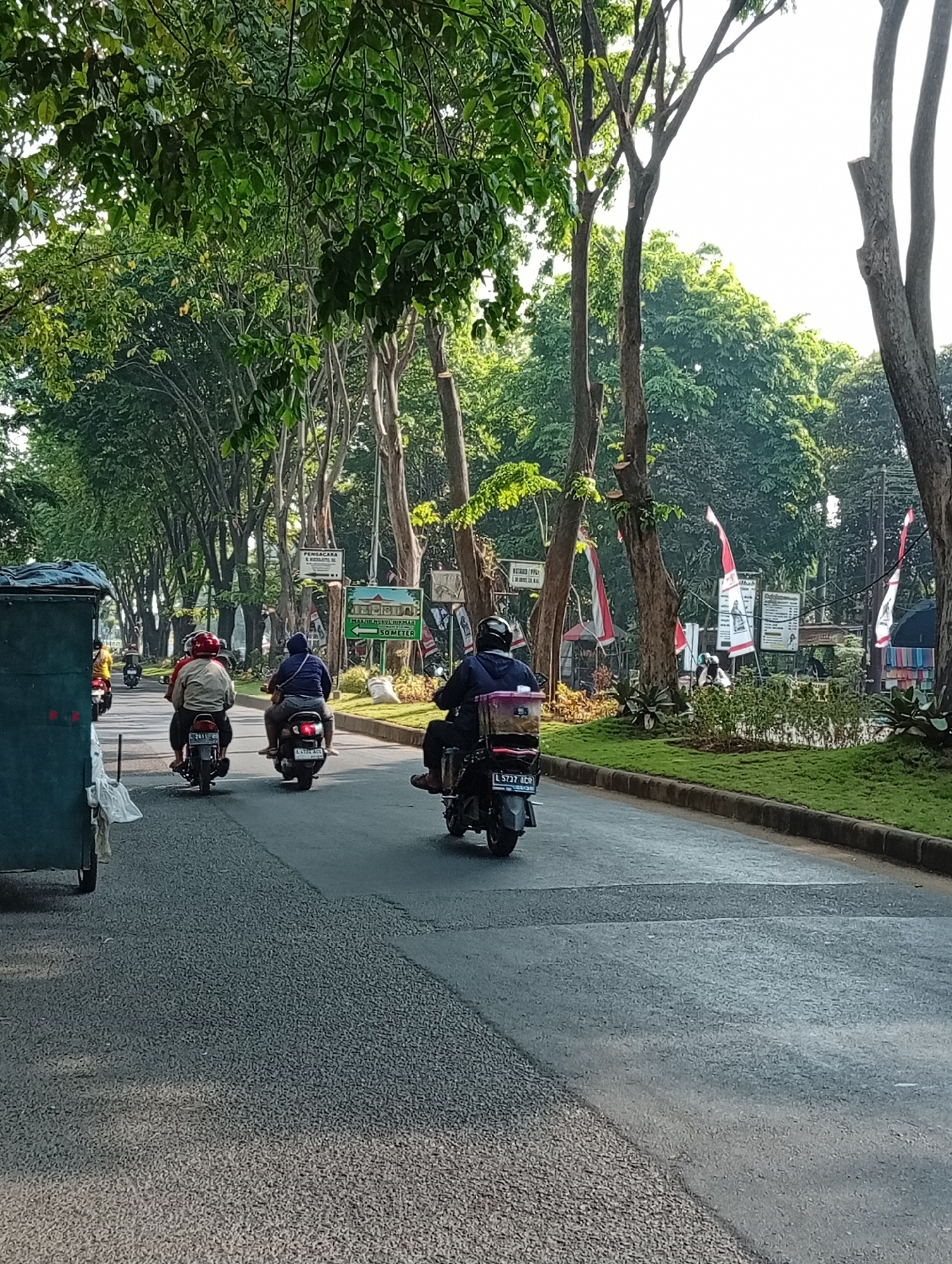 A street scene in Sepande, Indonesia, shows several motorbikes traveling down a road lined with trees. On the left, a teal cart is parked. Two motorbikes are in the foreground, one with two riders and the other with a single rider. A third motorbike is further down the road, with a single rider. Signs and red and white flags are visible on the right side of the road, along with neatly manicured grass. The sky is bright and appears to be daytime.