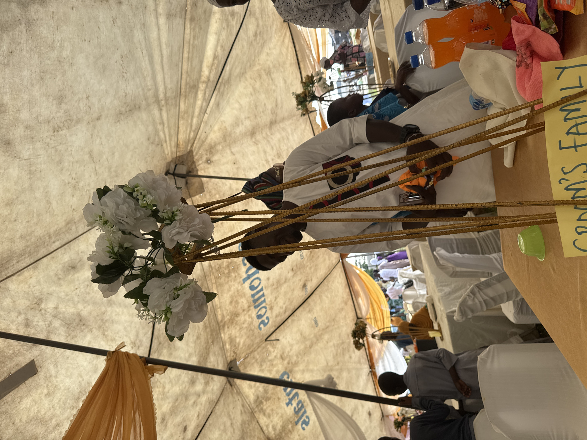 This image depicts an outdoor gathering, likely a celebratory event, under a large, tan-colored tent structure. The tent appears to be a temporary setup, possibly for a wedding or festival, given the presence of decorated tables and seated guests. In the foreground, a table is covered with an orange tablecloth, and a yellow sign with blue writing that says "Groom's Family" is visible. Several bottles of orange soda and a small green bowl are also on the table, along with folded cloths in pink and white.
A man wearing a white traditional Nigerian outfit, featuring intricate blue embroidery and a circular emblem, stands prominently in the mid-ground. He is holding an orange cloth and appears to be engaged in an activity related to the event. Several other individuals are visible in the background, some seated at tables draped with white tablecloths, while others are standing. The seating arrangement suggests a meal or reception is in progress. The overall environment is bright, indicating daylight. The distinctive blue lettering on the tent's fabric, which reads "Café" and "Somob", suggests a potential connection to a local establishment in Badagry, Nigeria, known for its coastal location and historical significance. The presence of the tent, the seated guests, and the decorative elements point to a cultural event being held in this Nigerian city.