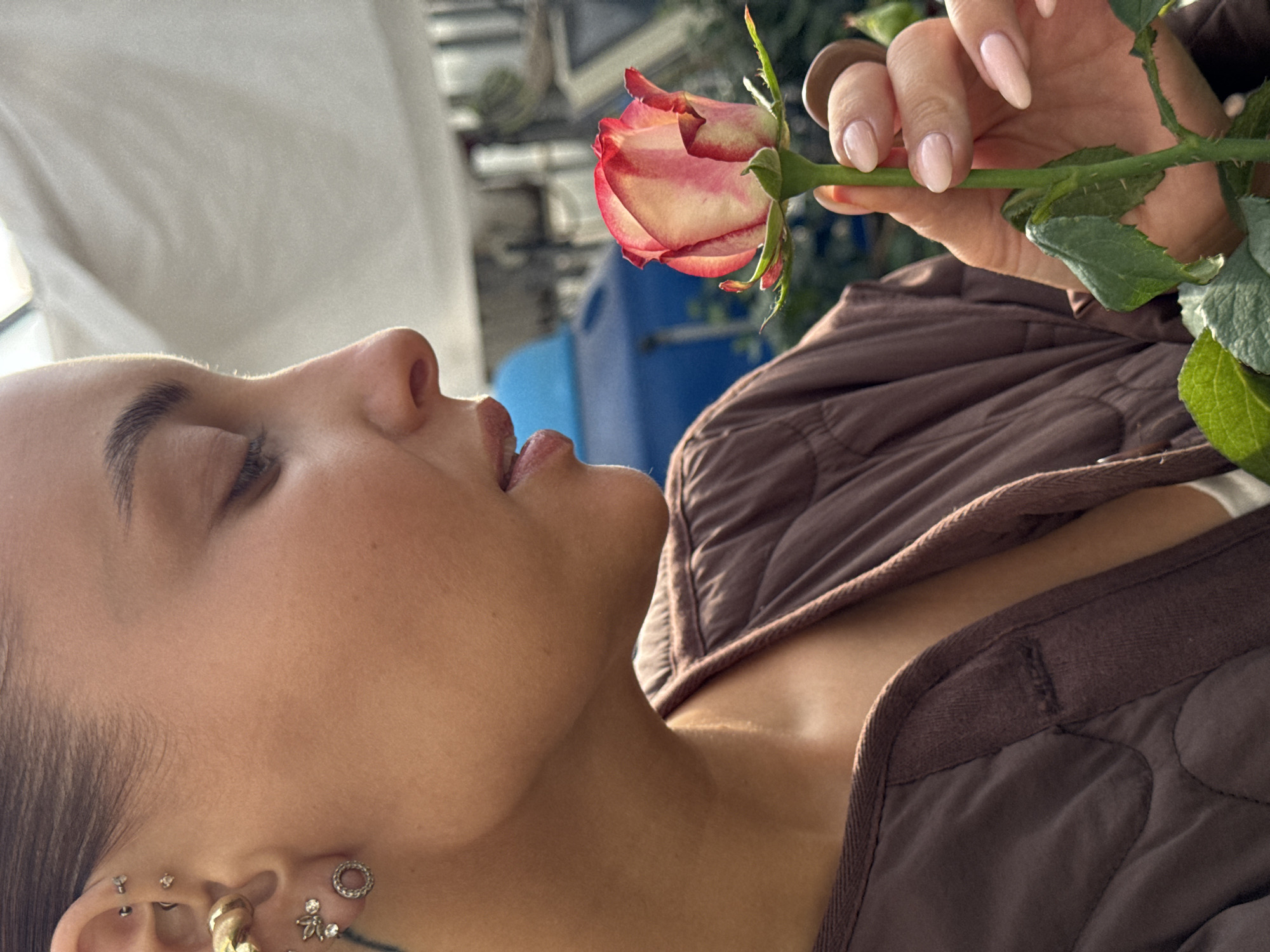 A woman with dark hair pulled back is shown in profile, smelling a single rose. She has several piercings on her ear, including a large gold hoop. She is wearing a brown quilted jacket. The rose is pink and white with green leaves. The background is blurred and appears to be outdoors with some greenery and a blue structure. The image does not contain any information to suggest the location is Chaikovsky, Russia.