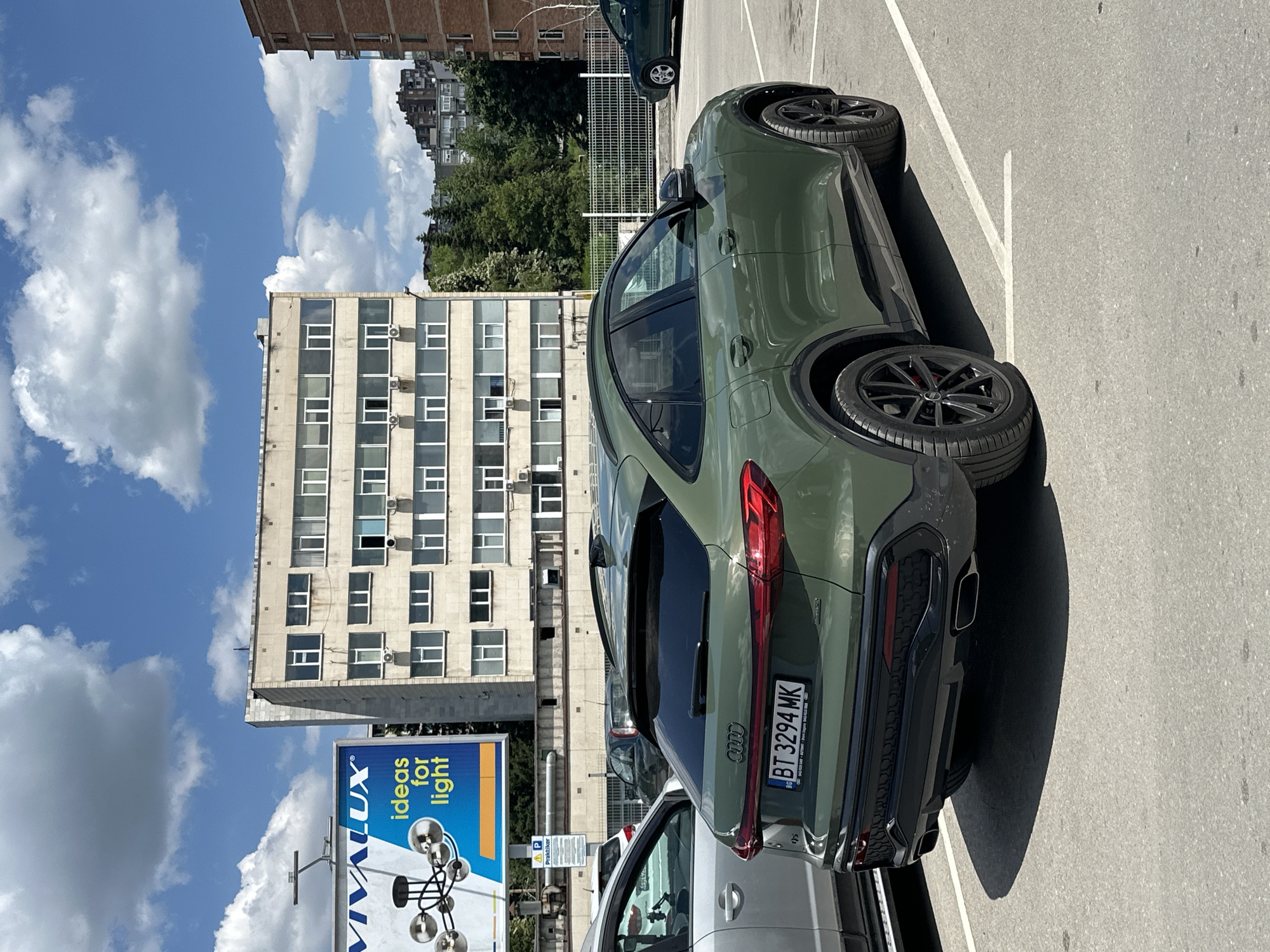 The image shows a dark green Audi Q5 parked in an outdoor parking lot on a sunny day with scattered clouds. The car is viewed from the rear, three-quarter angle. The license plate reads "BT 3294 MK". In the background, there is a multi-story building with numerous windows, a billboard advertising "VIVALUX ideas for light", and other buildings and trees. The parking lot is paved and marked with white lines. The scene appears to be in the city of Veliko Tarnovo, Bulgaria, as indicated by the "BT" prefix on the license plate, which is a Bulgarian code for Veliko Tarnovo. There are no people visible in the image.