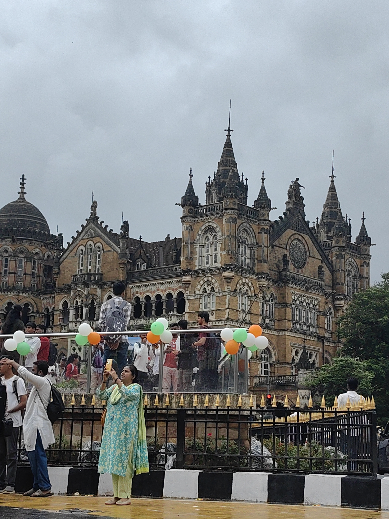 The image depicts the Chhatrapati Shivaji Maharaj Terminus in Mumbai, India, a UNESCO World Heritage Site. The grand Victorian Gothic architecture of the building is prominent, with its intricate stonework, multiple towers, and a large dome. A crowd of people is gathered in front of the terminus. In the foreground, a woman in a light blue and green patterned salwar kameez is taking a photo with her mobile phone. A man in a white kurta and blue jeans is also present, with his hand raised as if gesturing or shielding his eyes from the sky. Several balloons, in white, green, and orange, are tied to a railing, suggesting a celebratory occasion. The sky is overcast and cloudy. The ground appears wet, possibly from recent rain. Various individuals are visible in the background, some standing on an elevated platform.