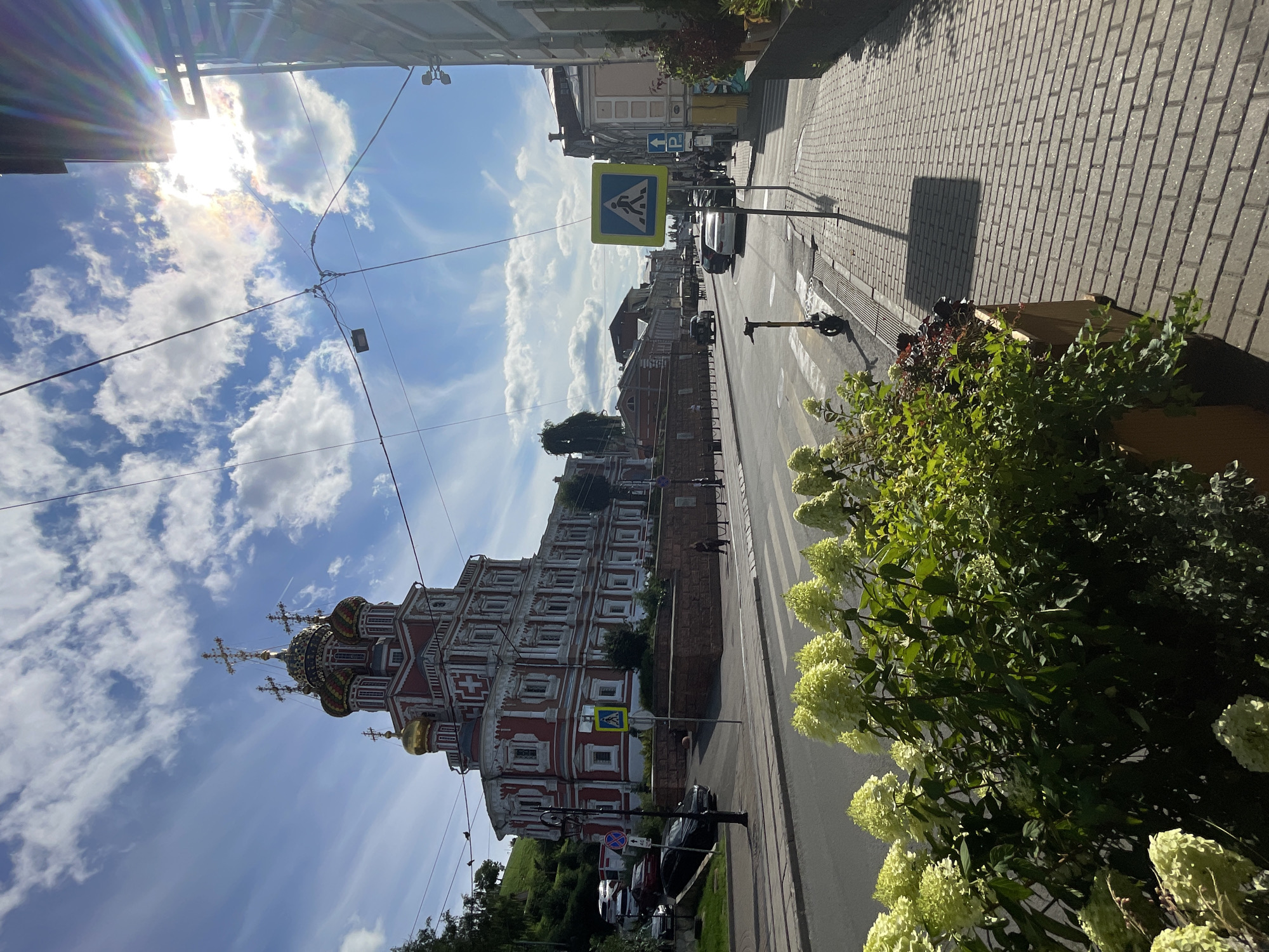 The image depicts a sunlit street scene in Nizhny Novgorod, Russia, with a prominent red and white church featuring golden domes on the left. The church, likely the Church of the Nativity of John the Baptist, is situated on a raised embankment. In the foreground, a paved sidewalk runs along the right side of the street, bordered by a planter box with light green hydrangeas. Across the street, there is a pedestrian crossing and an electric scooter parked nearby. Several cars are visible on the road, and buildings line the street in the background. The sky is blue with scattered white clouds, and the sun's rays are visible, creating lens flare. A pedestrian is walking on the sidewalk on the right, and a few people are visible further down the street near the buildings. Electrical wires are strung across the sky.