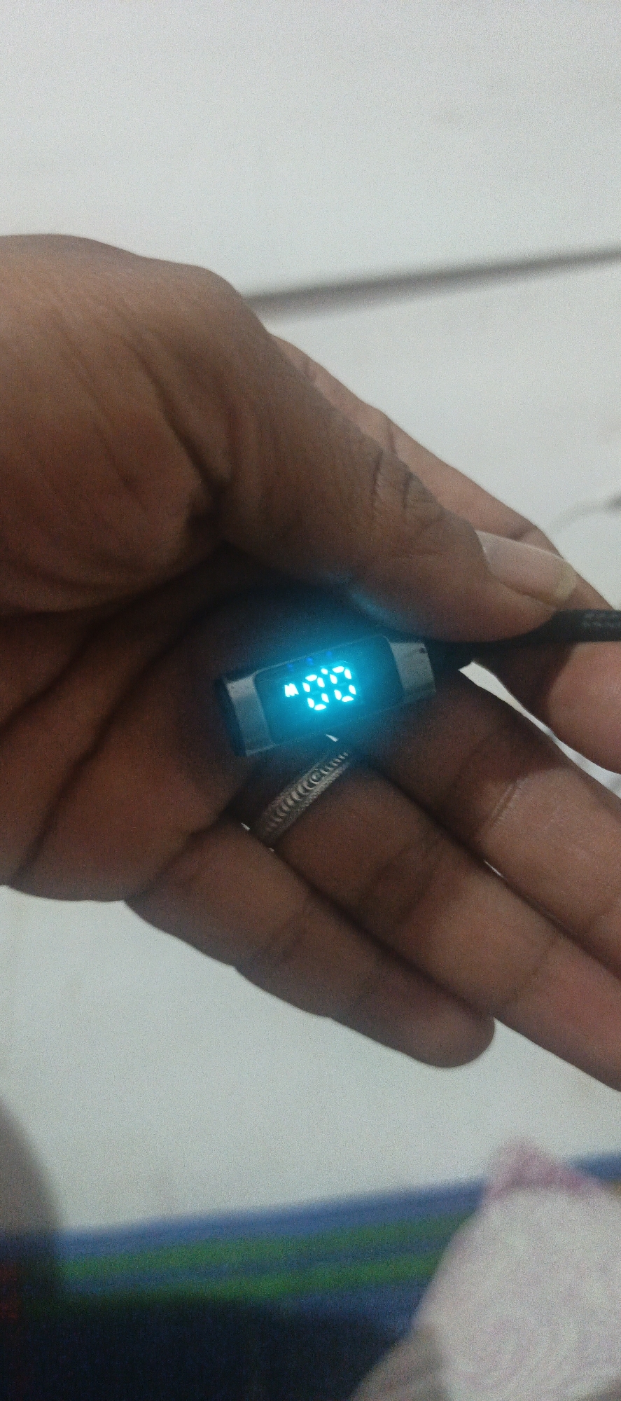 A close-up, vertical shot shows a hand holding a small, black electronic device with a bright blue digital display. The display shows "00" in large, blocky digits, with a small "A" to the left of the numbers. The device appears to be a USB tester or charger indicator. The hand is brown-skinned and appears to belong to an adult. A silver-toned ring with an etched pattern is visible on the ring finger. The background is a blurry, light-colored surface, possibly a table or wall, with some indistinct colored lines in the lower portion of the frame. The image is underexposed in some areas. There is no discernible information in the image to suggest it is located in Cinta Dame, Indonesia.