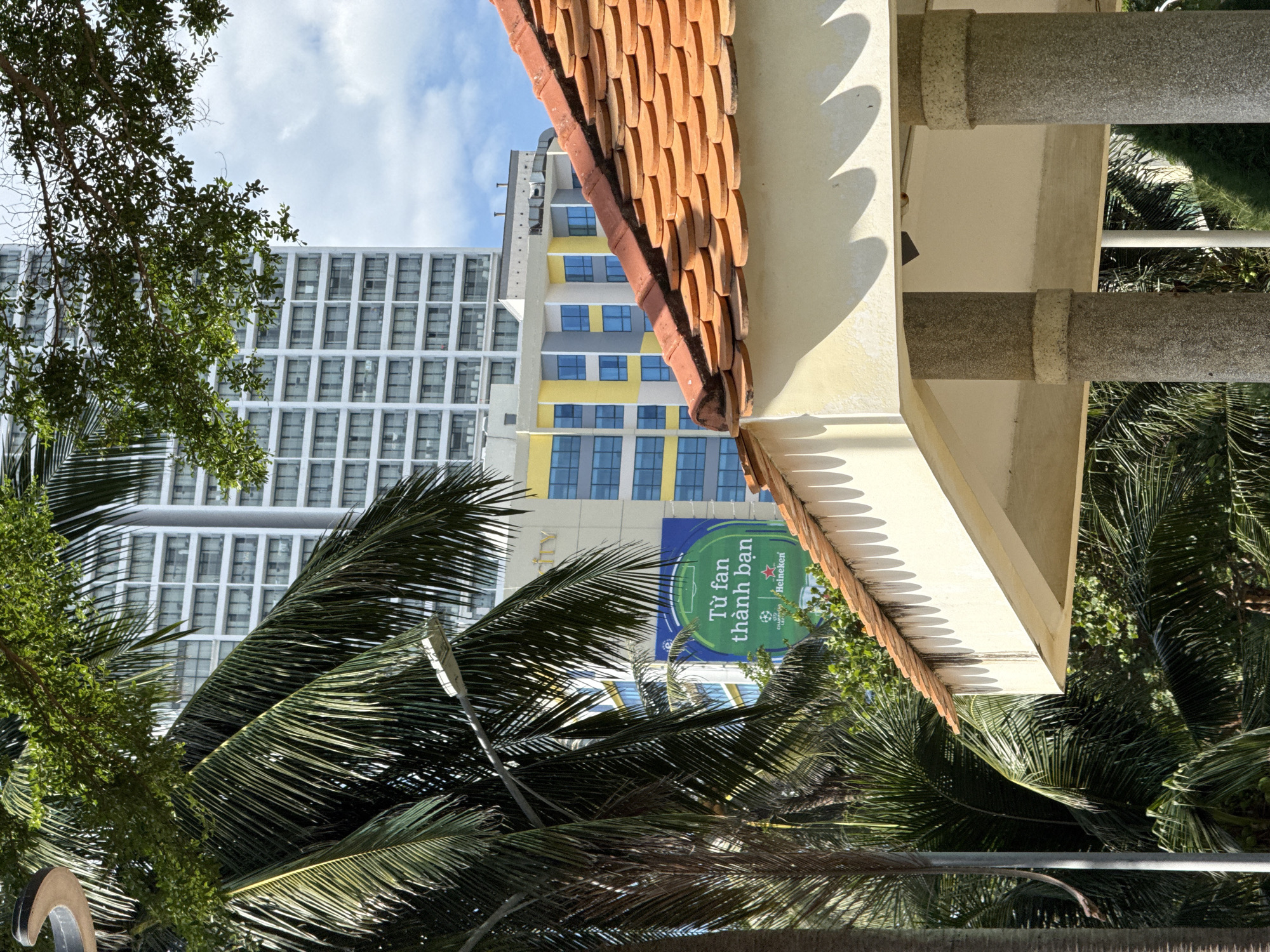 This image captures a scene in Nha Trang, Vietnam, featuring a prominent building with a distinctive tiled roof in the foreground. The building is tall and modern, with numerous windows, and appears to be a hotel or residential complex. In front of it, a large banner is displayed, with text in Vietnamese that translates to "From fan to friend." This banner also features the Heineken logo, suggesting a sports-related promotion.
The foreground is dominated by the tiled roof, which has an orange-brown hue and is laid in a pattern of overlapping semi-circular tiles. Supporting the roof are two large, cylindrical columns constructed from speckled stone. Palm trees with lush green fronds are visible on the left side of the image, partially obscuring the view of the buildings and banner. One of the palm trees has a streetlight attached to its trunk. The background shows a clear blue sky with some clouds. The overall impression is a sunny day in a tropical urban environment.