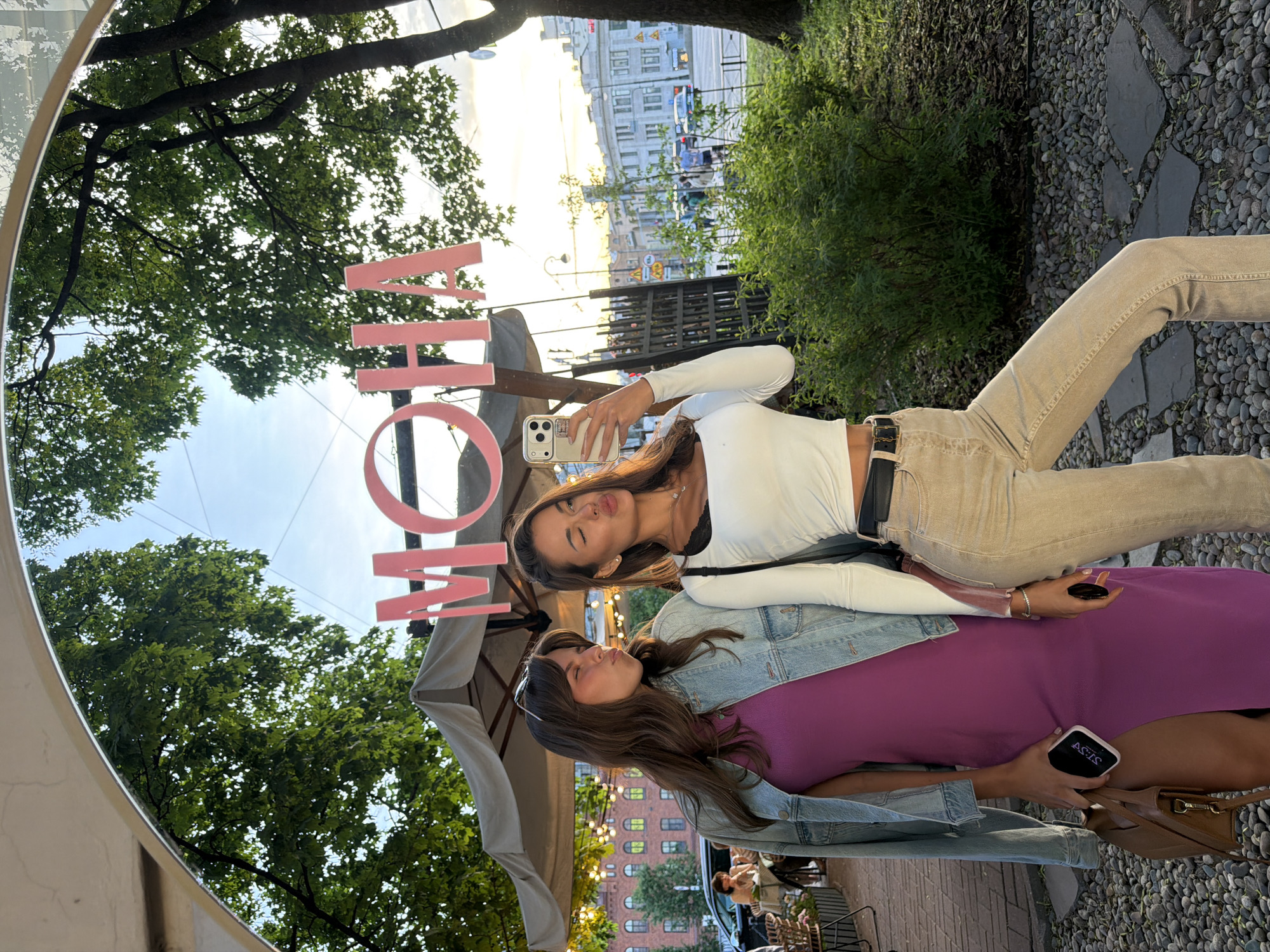 Two women are posing for a selfie in front of a mirrored surface that reflects a street scene in Saint Petersburg, Russia. The mirrored surface has large pink letters that spell out "MOHA". The woman on the left is wearing a purple dress and a denim jacket, and is holding a phone in her left hand. The woman on the right is wearing a white crop top and beige pants, and is holding a phone in her right hand. Both women are puckering their lips for the photo. In the reflection, trees, buildings, and a street with cars can be seen. The lighting suggests it is late afternoon or early evening.