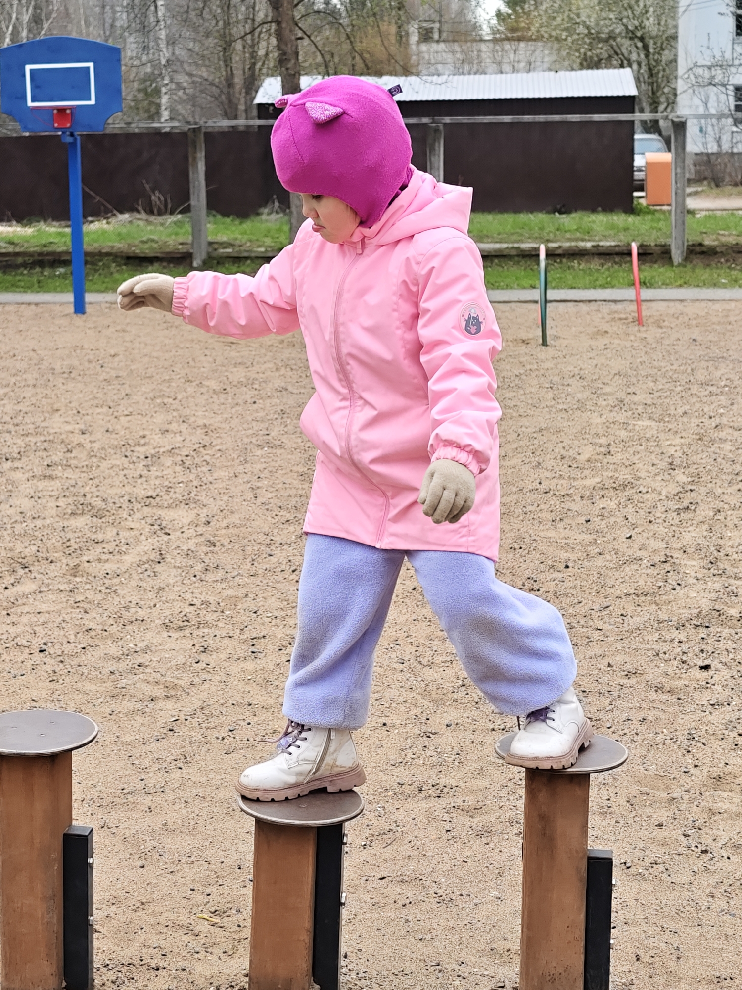 A young child, likely a girl, is the central figure in this outdoor image, captured in what appears to be a playground or recreational area. The child is dressed for cool weather, wearing a vibrant pink hooded jacket, a matching pink hat with small ear-like protrusions, light purple fleece trousers, and light-colored boots with brown soles and purple laces. She also has light beige gloves on both hands.
The child is actively engaged in balancing, carefully stepping across two brown posts topped with flat circular platforms. Her arms are slightly extended to the sides, indicating concentration and an effort to maintain balance. Her facial expression shows focus.
In the background, the scene reveals several elements: a blue basketball hoop with a white backboard stands to the left. Further back, a wooden fence separates the play area from what appears to be residential or utility buildings, one with a corrugated metal roof and another white structure. Bare trees are visible, suggesting either early spring or late autumn. Two colorful poles, one red and one blue/green, are partially visible sticking out of the sandy ground. The ground itself is composed of sand or fine gravel.
The lighting is natural and appears overcast, creating soft, diffused light without harsh shadows, consistent with the cool weather indicated by the child's attire. On the left sleeve of the child's pink jacket, a circular patch featuring a grey animal head (possibly a cat or wolf) inside a rainbow emblem is visible. The overall setting points to an outdoor play area in Chaikovsky, Russia.