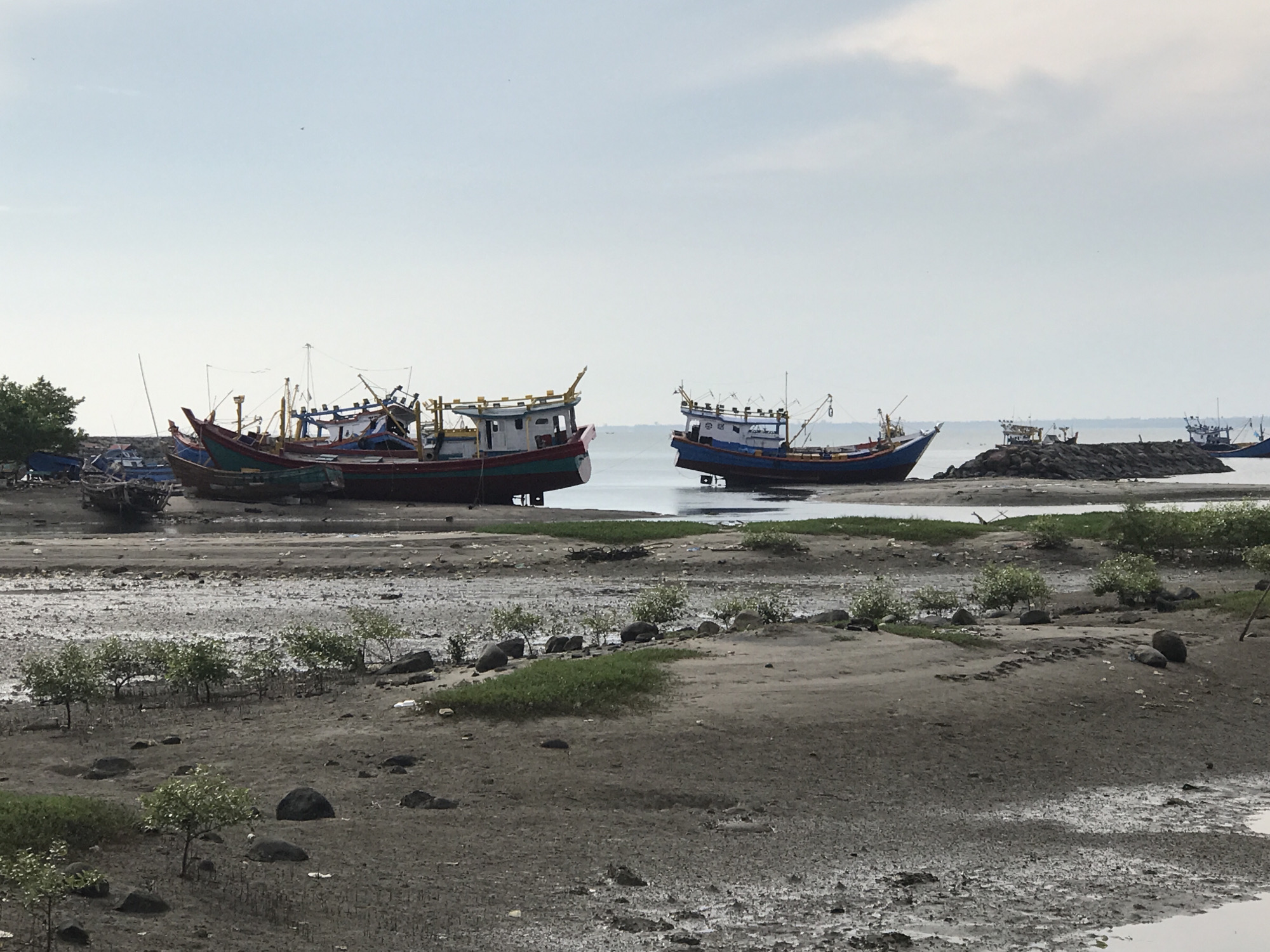 The image depicts a coastal scene in Lhokseumawe, Indonesia, with several fishing boats moored on a muddy shoreline. The sky is overcast with scattered clouds, and the water appears calm. The foreground is dominated by a muddy, tidal flat with sparse vegetation and scattered rocks. In the middle ground, a collection of colorful fishing boats, primarily red and blue, are beached or anchored in shallow water. These boats vary in size and condition, with some appearing weathered and others more recently maintained. In the background, the sea stretches to the horizon, with faint outlines of more boats and land visible. The overall impression is one of a working harbor or a place where fishing vessels are docked.