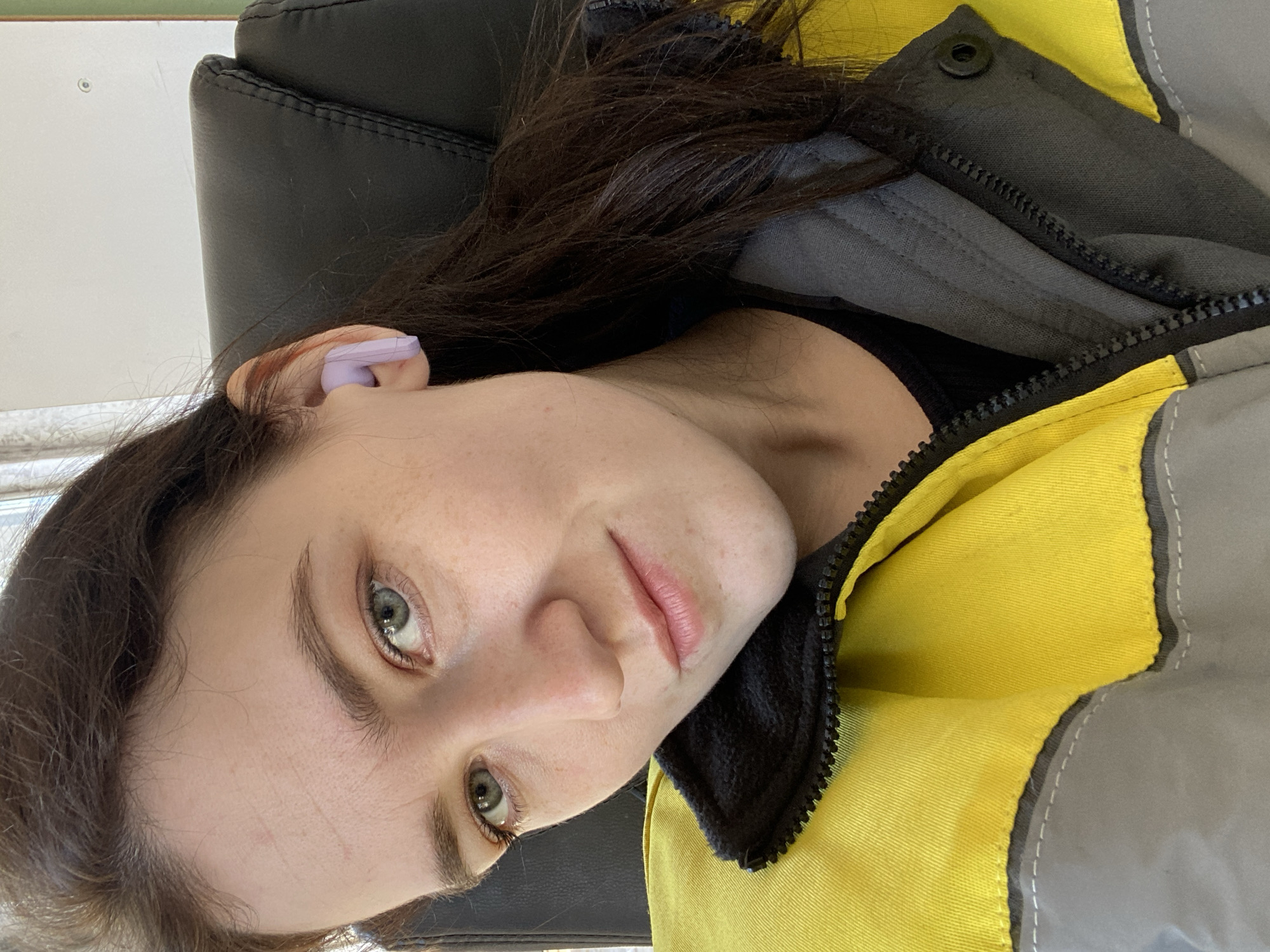 A close-up portrait of a young woman, rotated 90 degrees clockwise. She looks directly at the camera with light-colored (possibly blue-grey or green) eyes and a neutral to subtly content expression. Her light skin shows some natural freckling. Dark brown hair frames her face and flows downwards towards the bottom of the frame (due to the rotation).
In her visible ear, positioned towards the top left of the rotated image, she wears a light purple earbud. She is dressed in a sturdy-looking jacket, characterized by large sections of bright yellow and grey fabric, accented with black zippers and a black fleece collar. A black snap button is visible on the grey section near the zipper.
The background primarily features a black padded chair, seen behind her head and shoulder. A plain, light-colored wall or surface is visible to the upper left. The lighting appears soft and even, possibly natural light from an unseen window.
The primary activity is posing for the photograph, likely a selfie. There are no specific visual cues within the image to indicate the provided location context of Chaikovsky, Russia. No visible text is present.