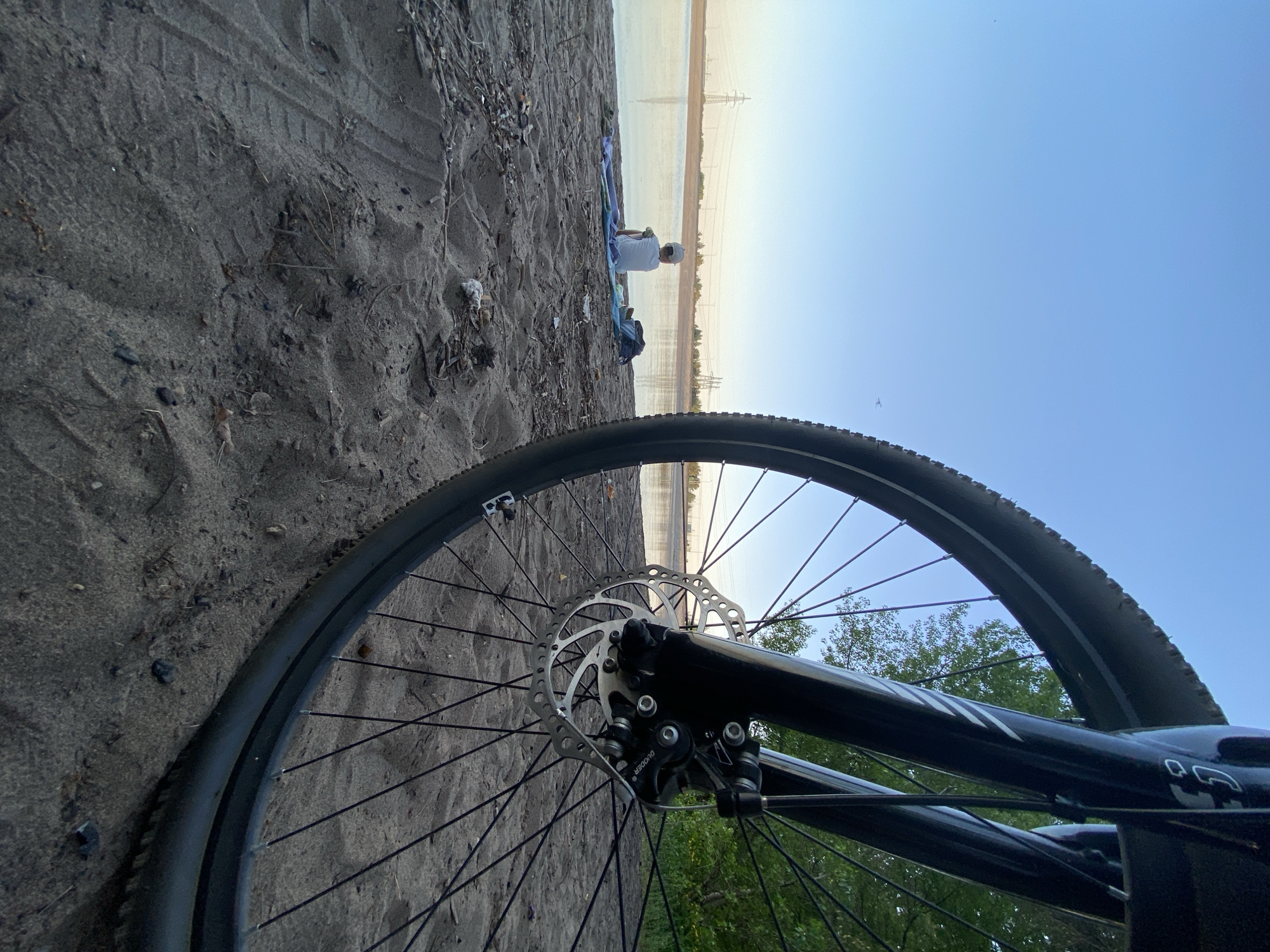 The image shows a bicycle wheel in the foreground, partially obscured by the sandy ground. In the background, a body of water is visible, with power lines spanning across it. On the shoreline, there is a person sitting on a blanket or towel. The sky is clear and blue. The location is in Noviy, Russia.