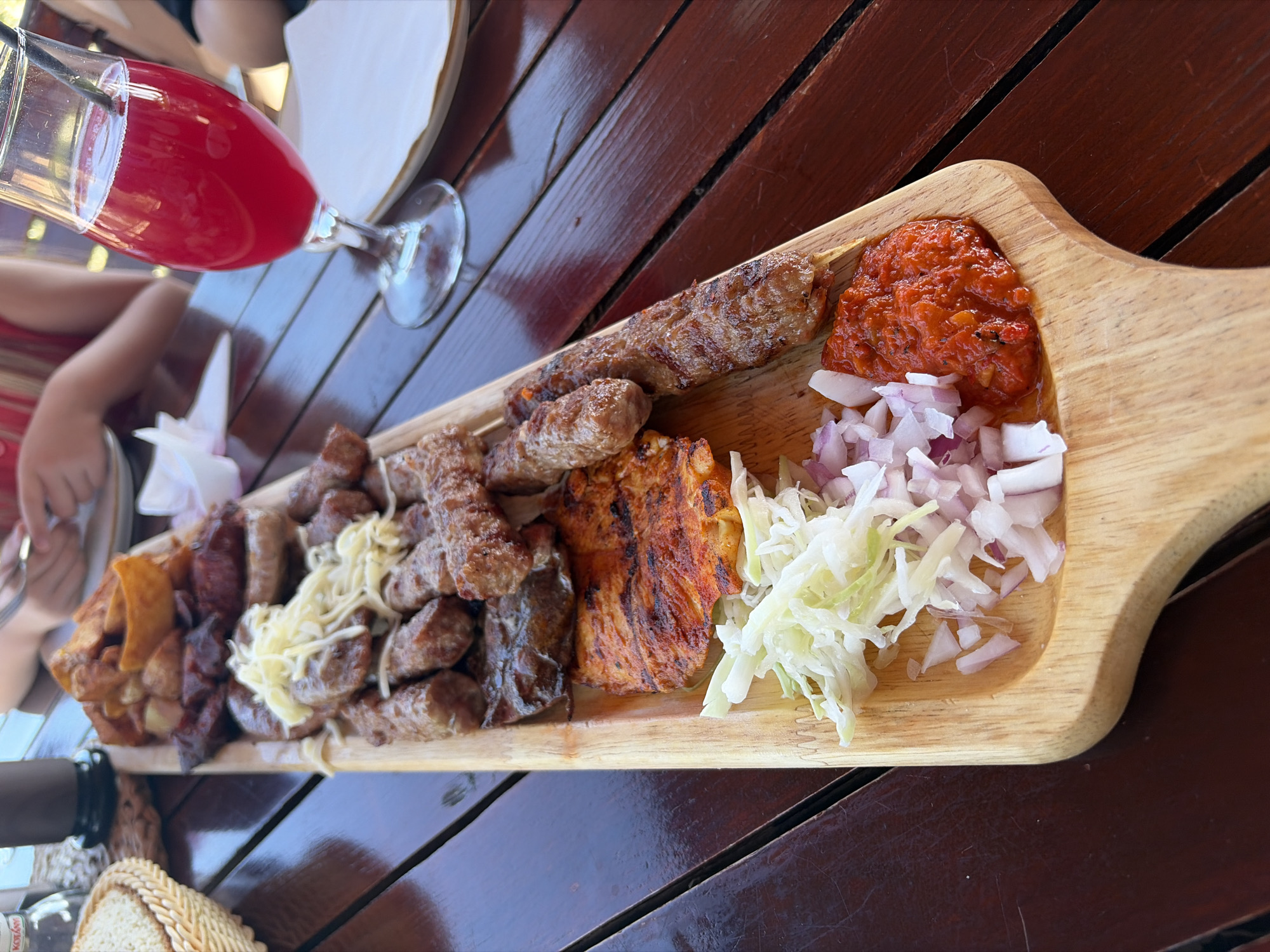 The image depicts an outdoor dining scene in Dobra Voda, Montenegro, featuring a large wooden platter of grilled meats and sides on a dark wooden plank table.
On the prominent light-colored wooden platter, a substantial assortment of grilled items is arranged. At the top, a pile of roasted or fried potato wedges is visible. Below this, various pieces of grilled meat include numerous small, elongated cylindrical items (likely ćevapi), several thicker grilled sausages, two longer pieces of grilled meat on skewers, and a large, flat piece of grilled meat or cheese with reddish seasoning and char marks. A mound of shredded, light-colored cheese or kajmak is placed over some of the ćevapi in the upper-middle section of the platter. At the bottom of the platter, from left to right, are distinct piles of shredded white cabbage, finely diced red onions, and a vibrant red relish or sauce (likely ajvar).
To the right of the wooden platter, a stemmed glass, characteristic of a wine or cocktail glass, holds a bright red/pink beverage with a dark straw protruding.
The table beneath the platter and drink is composed of dark brown wooden planks. In the upper left background, a portion of a woven basket is visible, along with a small, clear glass bottle and a dark-colored cup or container. Further in the background, out of focus, human hands are visible, one holding a fork over a white plate, indicating at least two individuals are present, one possibly wearing a red or striped garment. The overall lighting suggests a bright, possibly sunny, outdoor environment.