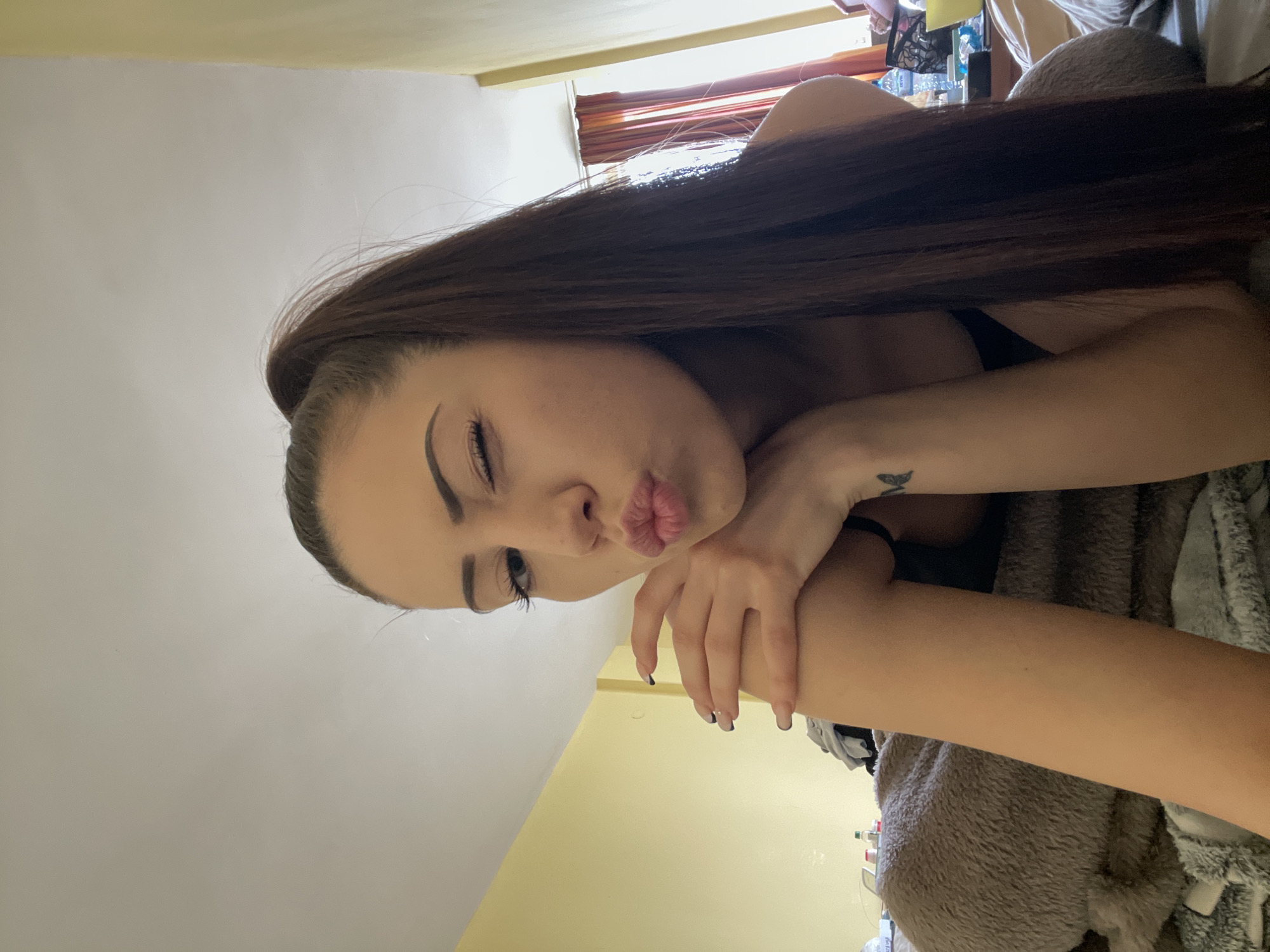 A woman with long brown hair is posing indoors. She has one eye closed and her lips pursed. A tattoo is visible on her left wrist. She is wearing a black top. The walls are painted yellow and white. A blanket is visible in the background. The location is unknown.