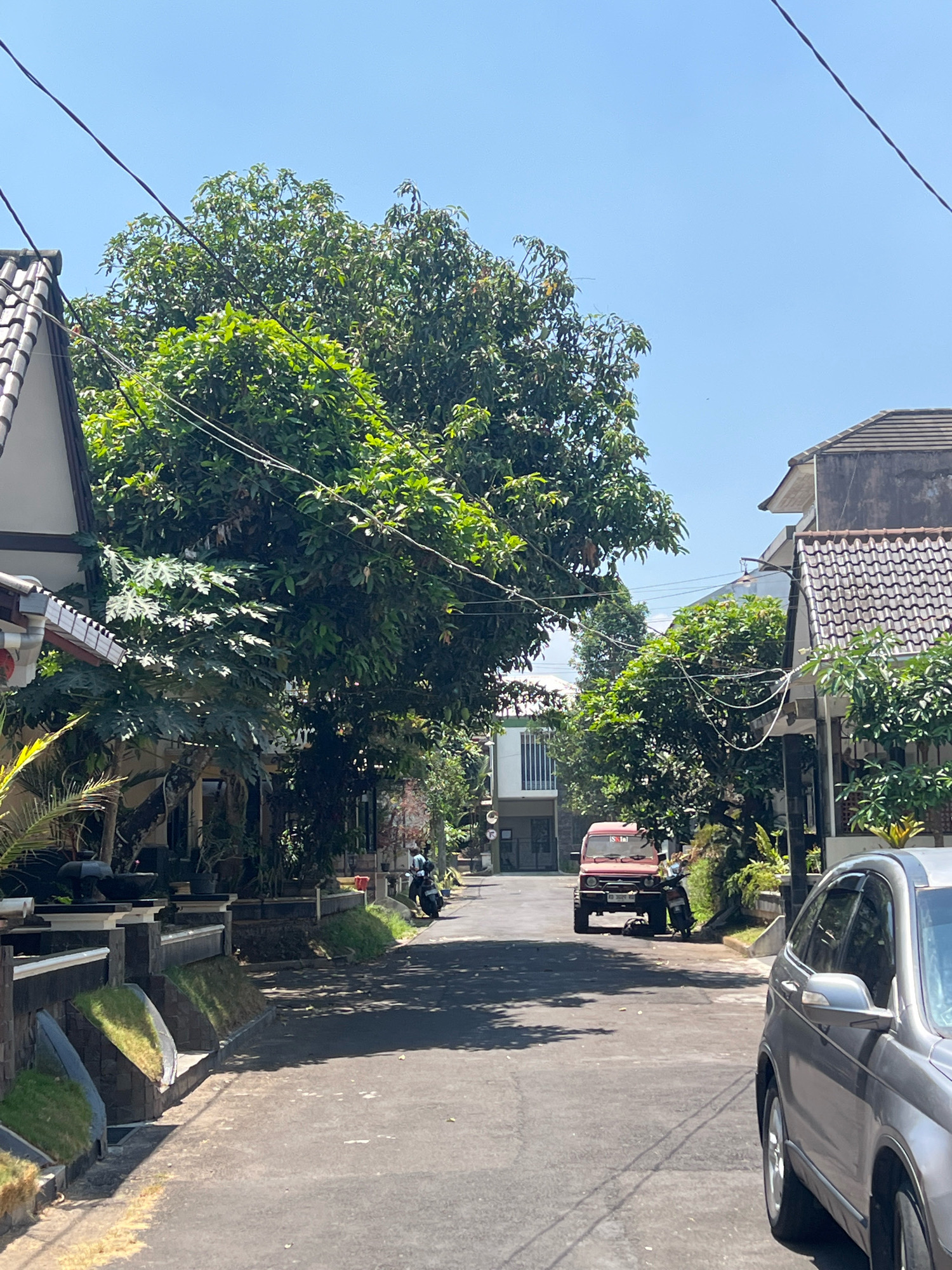 The image shows a residential street in Sleman, Indonesia, on a clear, sunny day. The street is paved and lined with houses and trees. In the foreground, the right side of a silver SUV is visible, parked or moving slowly on the right side of the road. Further down the street, a red SUV, possibly a Suzuki Jimny, is parked on the left side of the road, facing the camera. A black motorcycle is parked to the right of the red SUV. Tall green trees with dense foliage overhang the street, casting shadows on the asphalt. The houses on either side of the street have tiled roofs and are partially obscured by the trees and vegetation. The sky is a bright, clear blue. The overall impression is a quiet, residential neighborhood on a warm day.