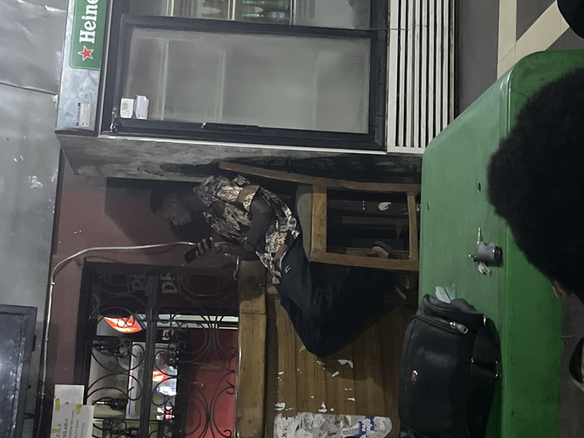 In Gura Top, Nigeria, an outdoor establishment is depicted at night. A young man sits on a wooden chair, engrossed in his mobile phone, wearing a patterned shirt and dark trousers. Behind him, a Heineken branded refrigerator stands tall, its glass doors reflecting the ambient light. A counter runs along the background, adorned with metalwork and glimpses of bottles. In the foreground, a green table occupies the space, holding a black bag and a set of keys. The general atmosphere suggests a casual gathering spot.
