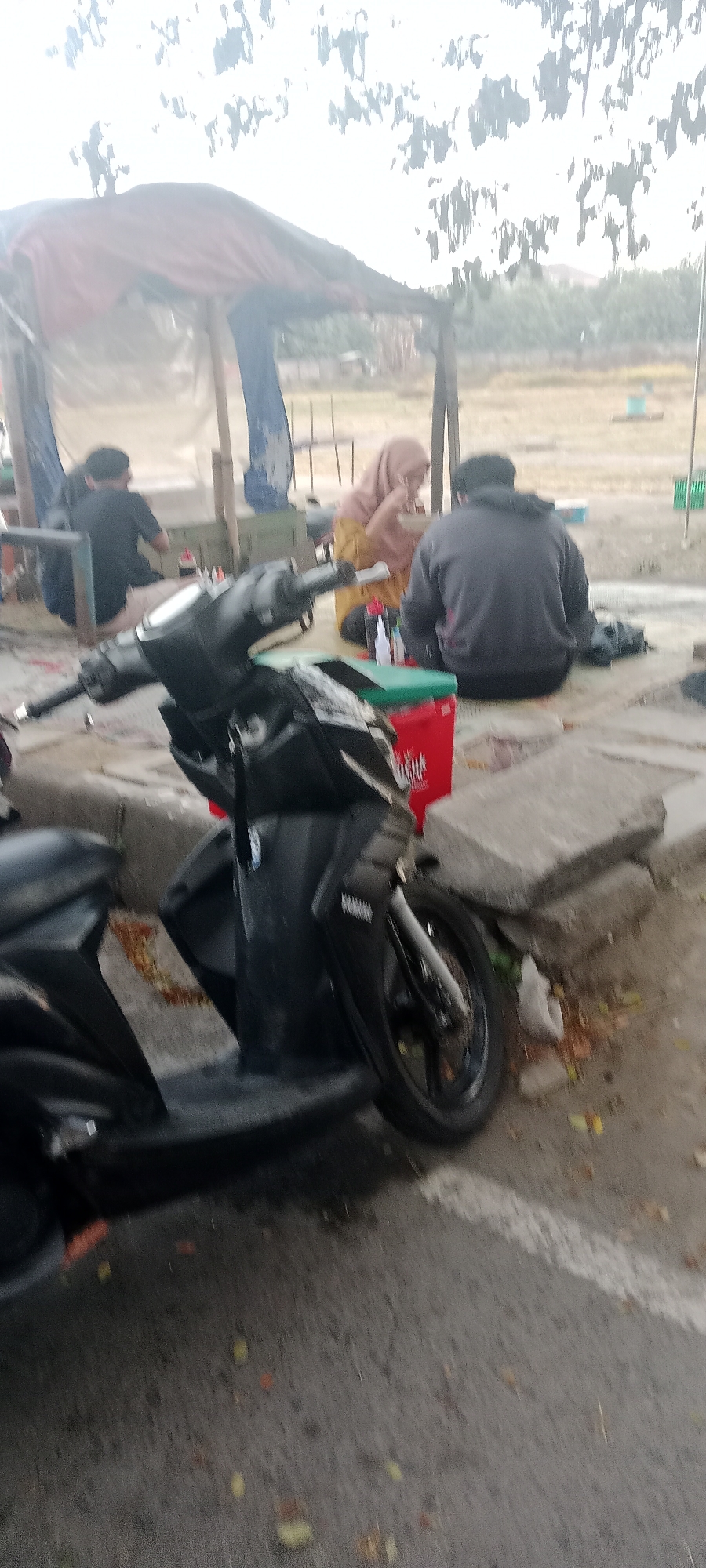 The image depicts an outdoor scene in Ngemplak, Indonesia, possibly a food stall or rest area. In the foreground, a black scooter is parked, partially obscuring the view of the subjects. Behind the scooter, three individuals are seated on the ground. To the left, a person wearing a black t-shirt is facing away from the camera. In the center, a woman in a light brown hijab and yellow clothing is eating from a bowl. To the right, a person in a dark grey hooded sweatshirt is also facing away from the camera. They appear to be in a casual setting, with a red cooler bag and some bottles on a green surface nearby. The background shows a makeshift shelter with a brown tarp and blue fabric, suggesting a temporary structure. The overall environment appears to be outdoors, with a blurred background of trees and vegetation. The lighting suggests it might be daytime, but the sky is overcast.