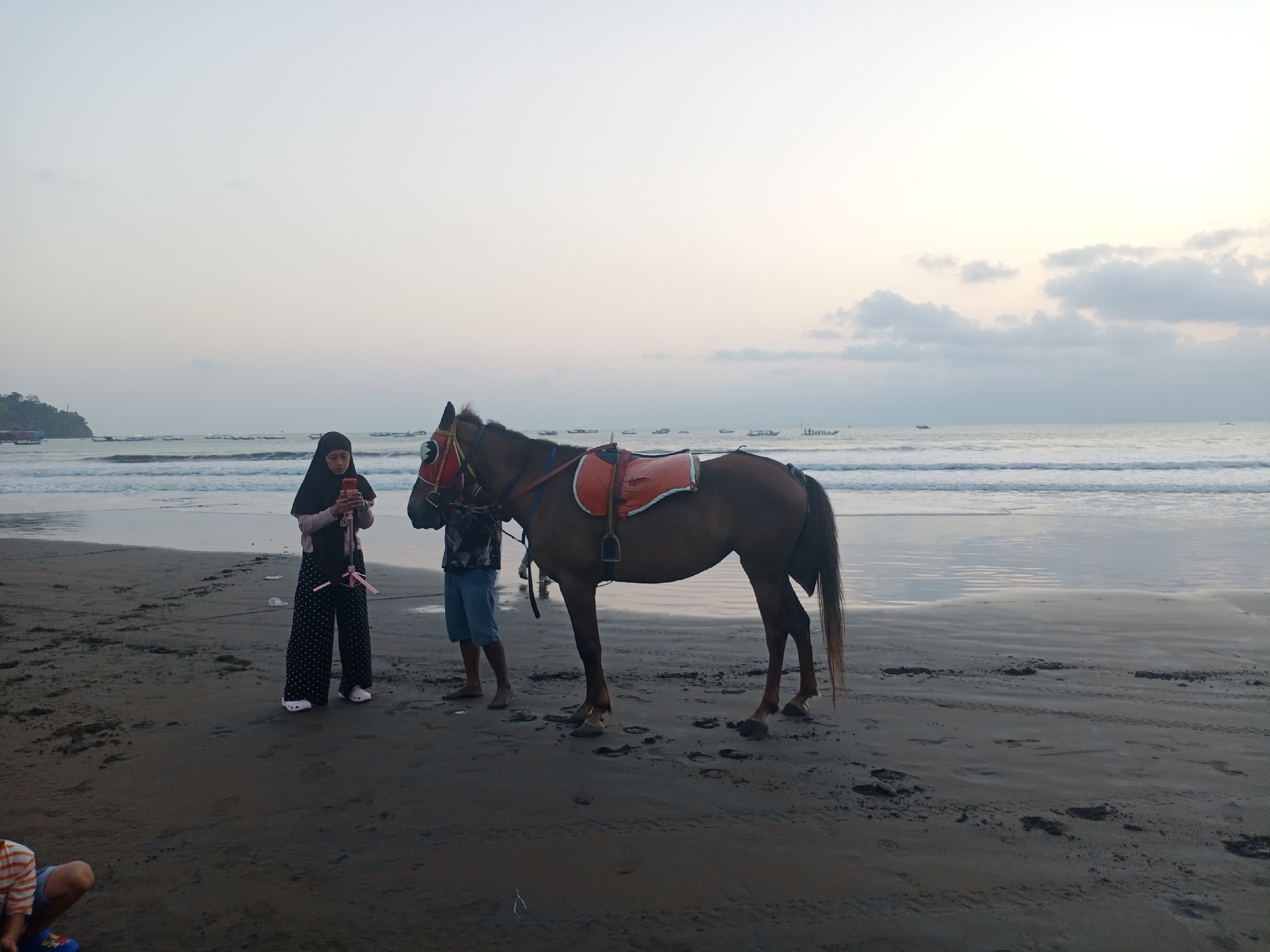 A brown horse is standing on a wet sandy beach in Desa Pagandaran, Indonesia. The horse is equipped with a saddle and reins. A person wearing a black hijab and patterned pants is standing to the left of the horse, looking at a mobile phone. A person wearing blue shorts is standing to the right of the horse. The ocean is visible in the background with small waves and several boats on the horizon. The sky is overcast with pale clouds. The sand on the beach has footprints and debris.