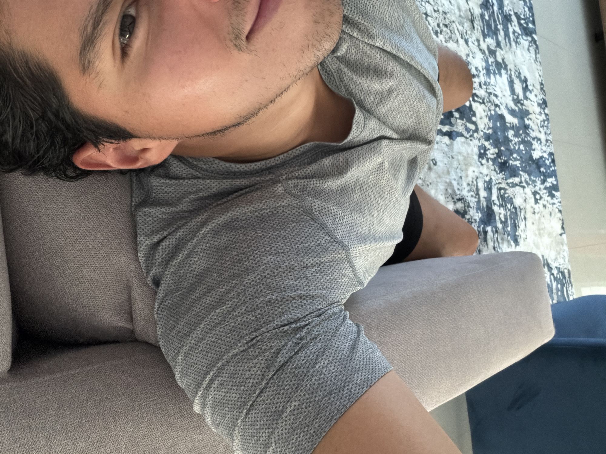 A man lies on a light gray couch. He is wearing a light gray T-shirt and black shorts. The background includes a white and blue rug, a tile floor, and a blue chair. There is no information in the image that indicates a specific location or any information about the city of Santa Clara Ocoyucan, Mexico.
