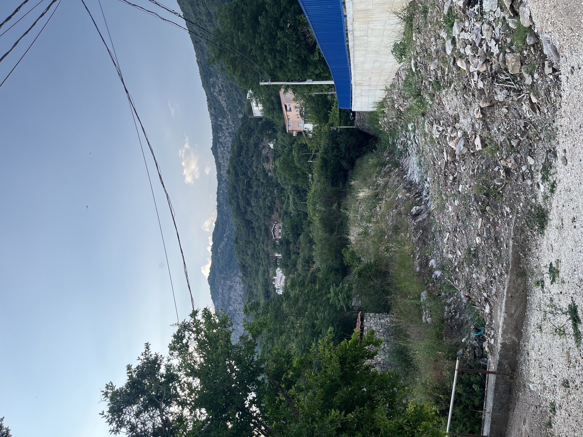 This media file depicts a rural hillside scene in Budva, Montenegro. The image is oriented vertically. In the foreground, the ground is covered in loose gravel and rocks, with some sparse green vegetation. A concrete pathway or retaining wall runs along the right side of the frame, disappearing into the distance. A metal pipe is visible, potentially part of a fence or structure.
Further up the hillside, dense green trees and foliage dominate the landscape, obscuring much of the terrain. Several houses or buildings are visible nestled within the greenery. One prominent building is a light pink, multi-story structure with a white trim and balconies. Another building below it appears to be white with red-tiled roofs. In the background, a steep, rocky mountain slope rises, covered in vegetation.
Above the hillside, a clear blue sky is visible with a few scattered white clouds. Several power lines stretch across the sky, intersecting the scene. A bright blue structure, possibly a roof or awning, is visible on the right side of the image, extending from a building or wall.
The lighting suggests it is late afternoon or early evening, with a soft light illuminating the scene. The overall atmosphere is peaceful and serene, capturing a glimpse of a residential area situated within a natural, mountainous environment. No people are visible in the frame, and no text is present.