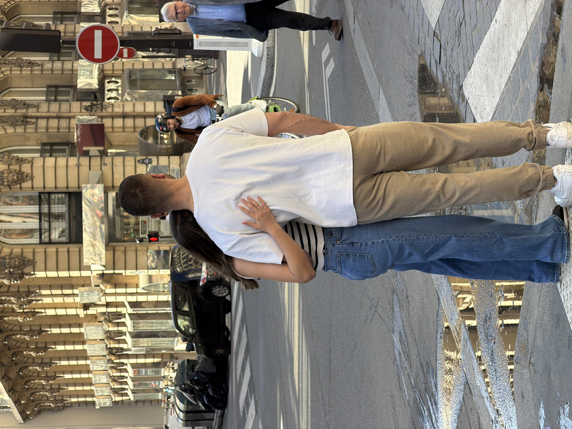 A couple is seen embracing on a street in Paris, France. The man has short dark hair and is wearing a white t-shirt and khaki pants. The woman has long dark hair, blue jeans, and a striped shirt. They are standing in the middle of a road with crosswalk markings, and a street with parked vehicles, and city buildings on the left side of the image. In the background, there are additional figures: a man in a suit walking near a "no entry" sign, and another person on a bicycle wearing a helmet. The pavement is visible, and there is architectural detail on the buildings lining the street.