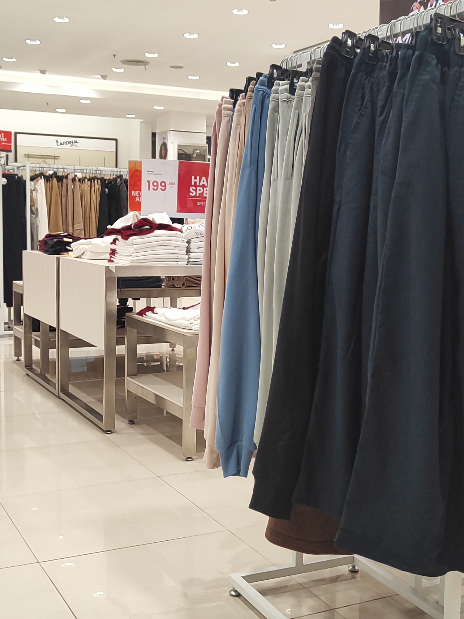 This image depicts a retail store interior, likely a clothing boutique or department store, with a focus on a rack of folded pants and several display tables. The pants on the rack are in various colors, including dark blue, grey, light blue, and pinkish-beige, and are neatly hung with black hangers. On the display tables, stacks of white and maroon folded shirts are visible. The store features bright overhead lighting and a tiled floor. A sign with "CARDINAL" branding is present in the background, suggesting the name of the store or a brand it carries. Another sign advertises a sale with prices like "199.900". The overall impression is one of a clean, organized retail environment. No people are clearly visible. The location is stated to be in Nangkaan, Indonesia.