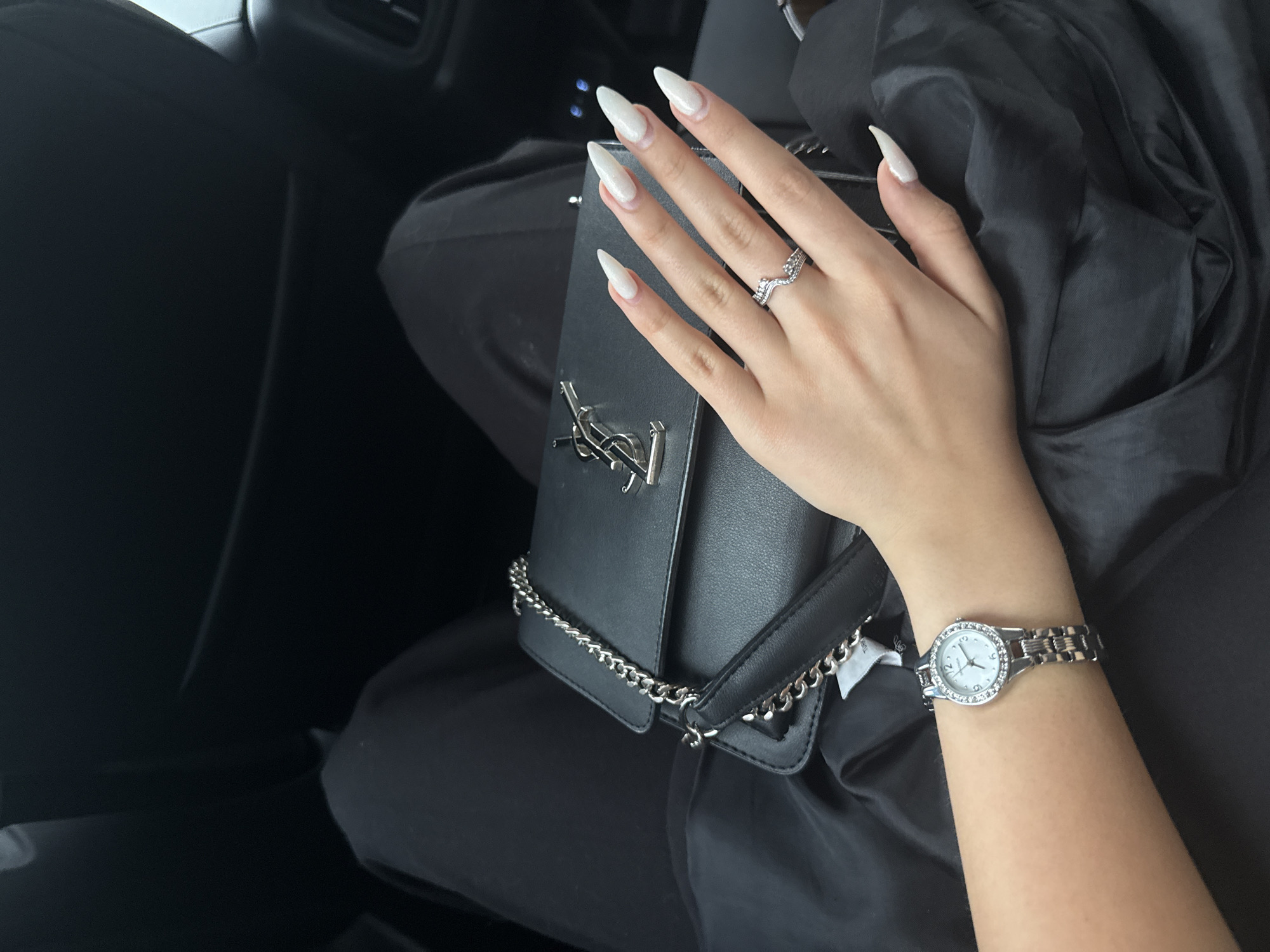 The image presents a close-up, first-person perspective view from within a vehicle, likely a car. The primary subject is a person's left arm and hand, resting upon a black shoulder bag situated on their lap. The hand features long, pointed, iridescent white artificial nails. A silver-toned ring with a wavy, chevron-like design, adorned with small rhinestones, is worn on the ring finger of the left hand. On the left wrist, a silver-toned watch with a round, light-colored face and a bezel encrusted with small rhinestones is visible; the word "QUARTZ" is discernible on the watch face.
The black handbag, appearing to be made of leather or faux leather, is embellished with a prominent silver-toned "YSL" (Yves Saint Laurent) logo on its front flap and has a silver-toned chain strap. Beneath the person's left arm and the handbag, a black fabric item, possibly clothing or a wrap, is visible, with a white tag partially showing the text "Turkey" or a similar word. The background consists of the dark interior of the vehicle, including a black car seat, a headrest, and a partial view of an air vent, consistent with a car's dashboard or door panel. The overall scene is static, with no discernible actions or interactions beyond the placement of objects. The environment is confined and interior. The specific location of Sofia, Bulgaria, cannot be ascertained from any visual cues within the image.