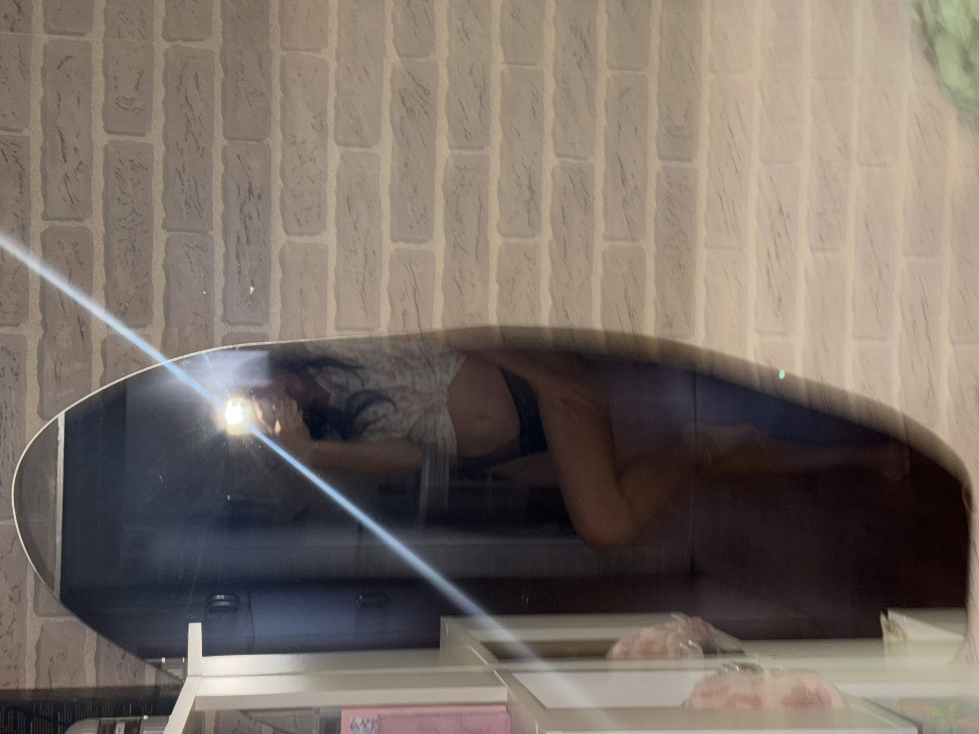 A full-length mirror reflects a young woman standing in front of a brick wall. She is wearing a light-colored, floral-patterned shirt tied at the waist, exposing her midriff, and dark underwear. Her right leg is bent at the knee, and she is holding a phone in her right hand, using it to take the photo. A bright light from the phone illuminates her face and upper body. The mirror is oval-shaped and reflects a shelving unit to the left, with some items on it. The background is a pale gray brick wall with visible grout lines. The image appears to have been taken indoors. The exact location in the city of Chaikovsky, Russia, is not discernible from the image.