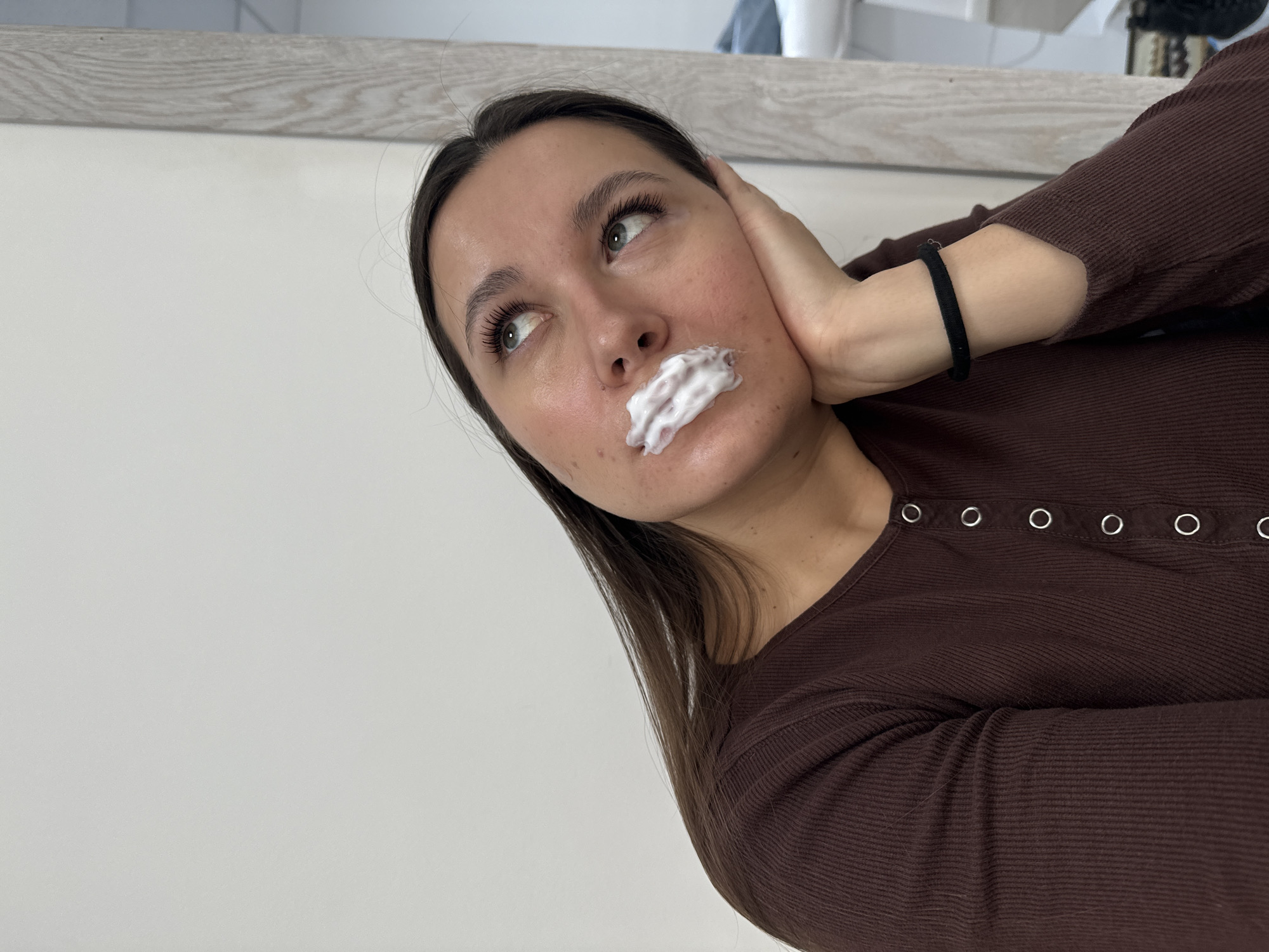 An adult female is depicted in an indoor setting, described as being in Odintsovo, Russia. She has long dark hair and is wearing a dark brown, ribbed long-sleeved top with a Henley-style neckline featuring multiple silver-rimmed buttons. Her head is tilted slightly back and to the viewer's right, with her eyes directed upwards. Her left hand supports her chin, and a black hair tie is present on her left wrist. A significant amount of white, foamy substance, consistent with a cosmetic treatment product, is applied to her upper lip, extending onto the philtrum and slightly below the nostrils. Her eyelashes appear long and dark, consistent with cosmetic enhancement. Visible blemishes are present on her chin and jawline. The background consists of a plain light-colored wall. A light-colored wooden door frame or partition is visible on the right side of the frame, behind which a blurred white object, consistent with a treatment bed or chair, and a dark object on the floor are partially visible.