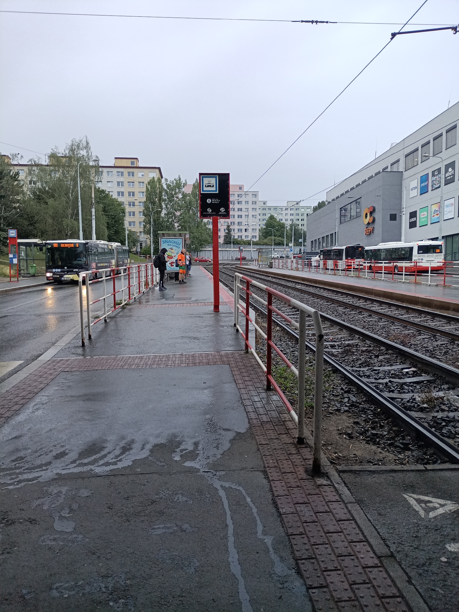 A bus and tram stop is depicted on a cloudy day in Prague, Czechia, likely near the "OC Repy" shopping center, indicated by signage. The scene is wet from rain, with puddles on the asphalt and tram tracks.
In the foreground, a wet, textured pavement leads to a raised platform with tactile paving. A red and white striped metal barrier separates the platform from the tram tracks. On the platform, several individuals are waiting, including a person in a black hoodie and backpack and another in a bright orange vest. They are positioned near a bus stop sign.
Behind the platform, tram tracks run parallel to the waiting area. Further back, a dark-colored bus with illuminated destination signs ("180 DEJVICE 6") is stopped at the bus stop. Beyond the bus, a large modern building complex is visible, featuring the "OC Repy" logo. Several shops, including "ACTION," "CREDO," "ALBER," "dm," and "LUXOR," are advertised on the building's facade.
In the distance, apartment buildings and trees line the horizon under an overcast sky. A red car is parked near the tram tracks in the background. The overall atmosphere is subdued due to the weather and the urban setting.