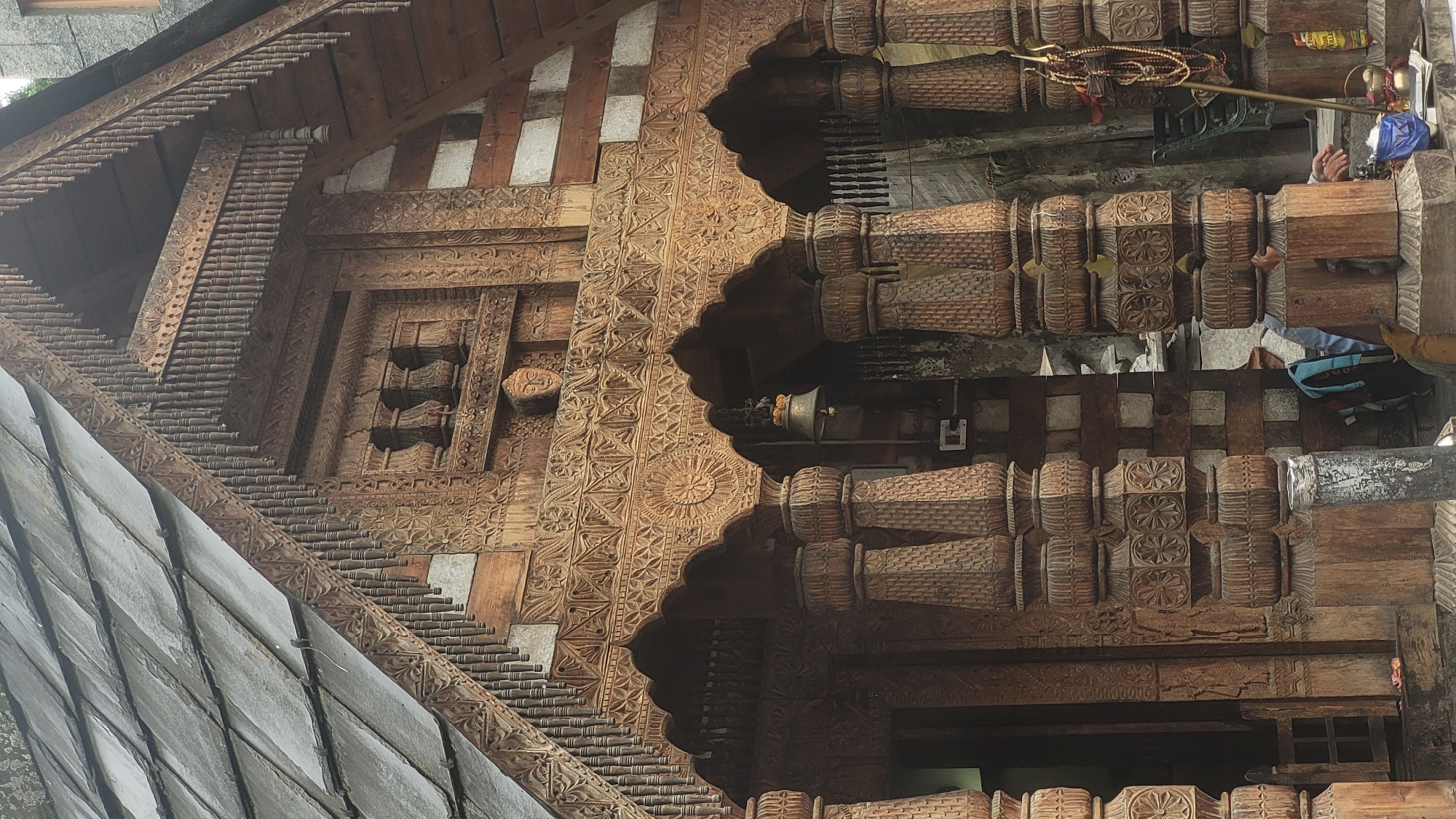 The image shows the intricately carved wooden facade of a building, likely a temple, in Vashisht, India. The architecture features traditional Himachali style with detailed woodwork. The roof is covered with grey slate tiles. Below the roofline, there is a large wooden frieze adorned with geometric patterns and floral motifs. Several arched openings are supported by elaborately carved wooden pillars, which exhibit stacked decorative elements and floral carvings. A bell hangs from one of the arches, and a carved wooden face is visible above the upper frieze. The building's walls appear to be constructed from stone and wood. In the foreground, a metal pole with prayer beads and a trident-like object is visible, suggesting a religious site. Two individuals are partially visible in the lower right portion of the image, one wearing a blue jacket and the other with hands visible. The overall scene depicts a culturally rich and architecturally significant structure in Vashisht.