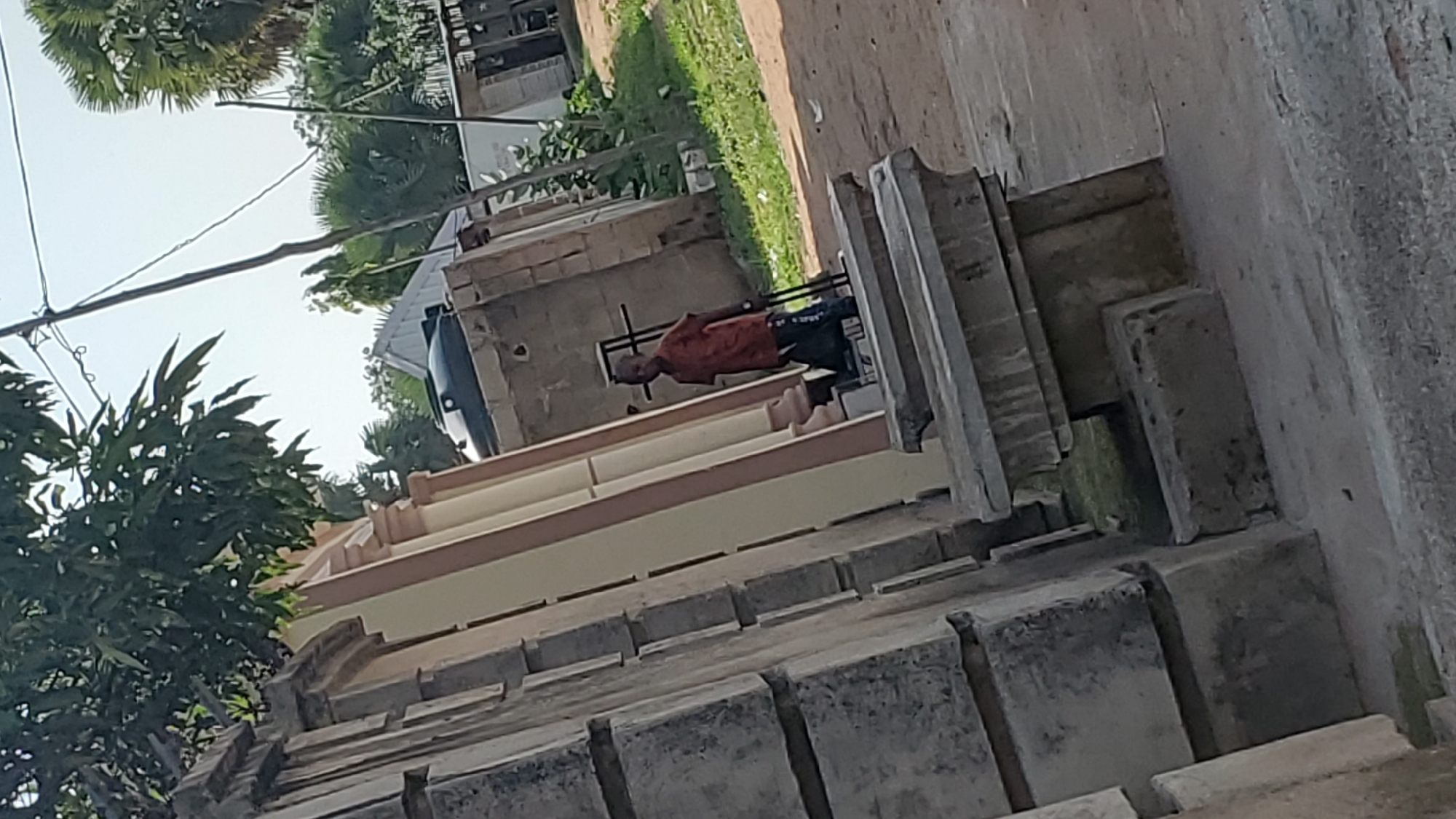 A young Black male stands in an outdoor environment with weathered, stone structures and vegetation. He is wearing an orange t-shirt and dark pants with white stripes. He is holding a black bag or object with a handle in his right hand. Behind him, there is a large black water tank and a sloped roof of a building. The background shows trees, a utility pole with wires, and a sandy ground. The lighting suggests it is daytime. The scene appears to be in a residential or communal area.