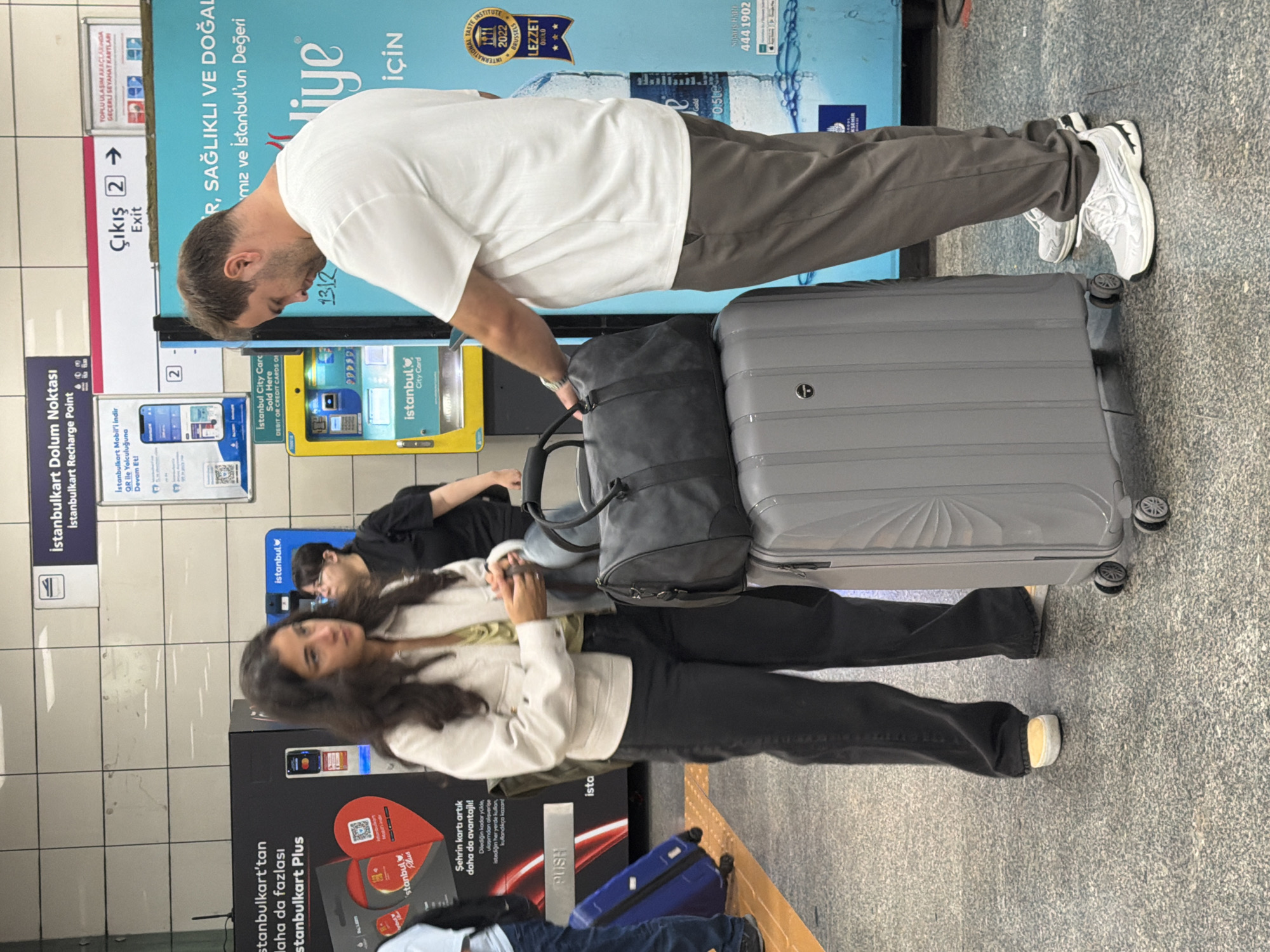 Two individuals, a woman and a man, stand near a luggage cart on a tiled floor. The woman, with long dark hair and wearing a light-colored jacket and dark pants, looks to her right, holding her hands together. The man, with short brown hair, wears a white t-shirt and grey pants. He is looking down and to his left. In front of them is a large grey suitcase, and on top of it rests a dark grey duffel bag. Behind them, a yellow vending machine is visible, and signs on the wall indicate "istanbulkart Dolum Noktası" (Istanbulkart Recharge Point) and "Çıkış 2 Exit" (Exit 2). The environment appears to be a public transport station.