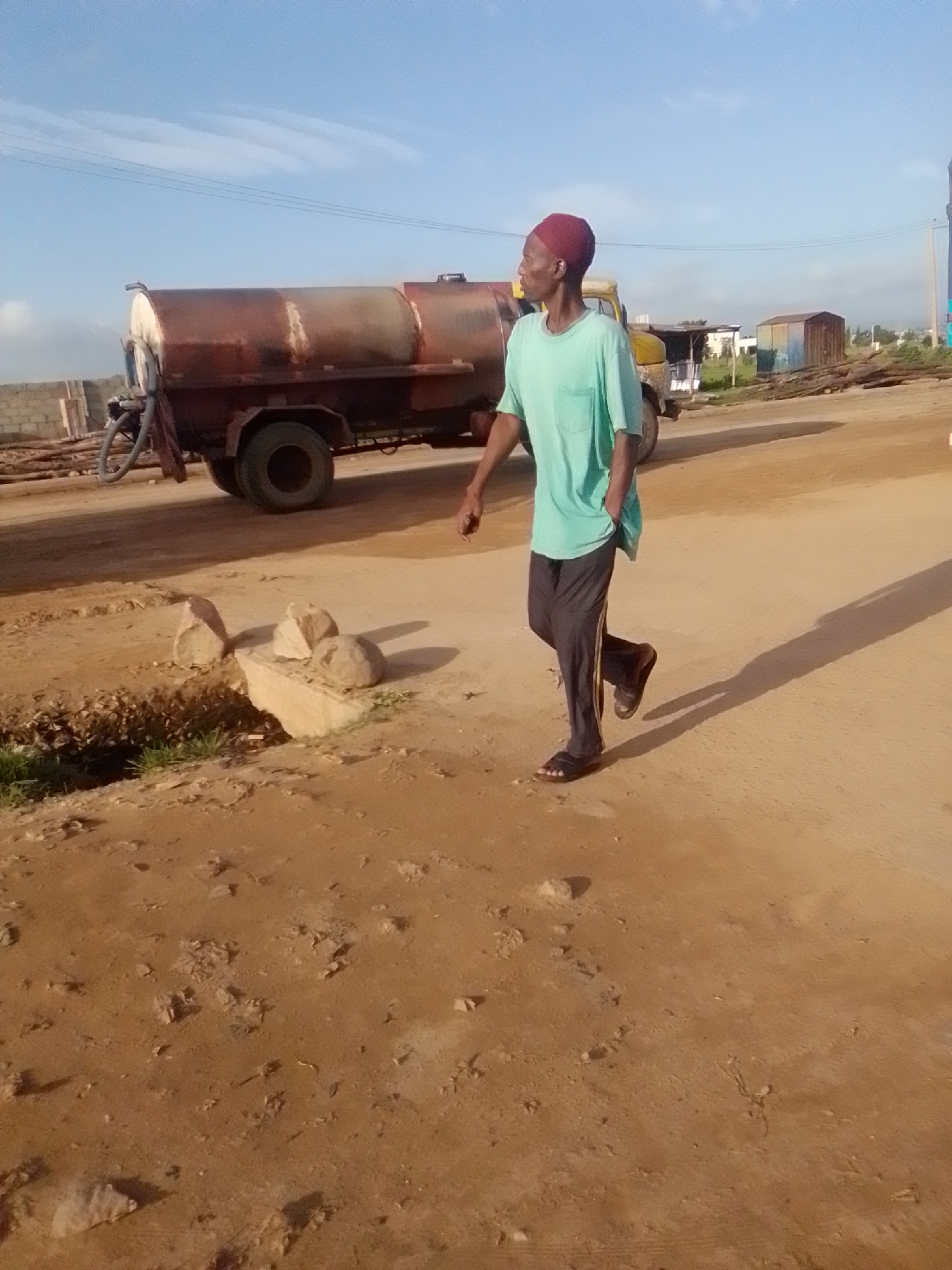In Kwadon, Nigeria, a man wearing a red cap and a light blue t-shirt walks along a dusty road next to a rusty water truck. The man has his left hand in his pocket and his right arm extended slightly. The truck, which appears to be a tanker, has two visible wheels and is parked on the side of the road. In the background, there are some buildings, including a small corrugated metal structure, and a clear blue sky with wispy clouds. The ground is a mix of dirt and scattered rocks, with some sparse vegetation visible. The overall impression is of an outdoor, likely industrial or construction site, setting in a sunny environment.
