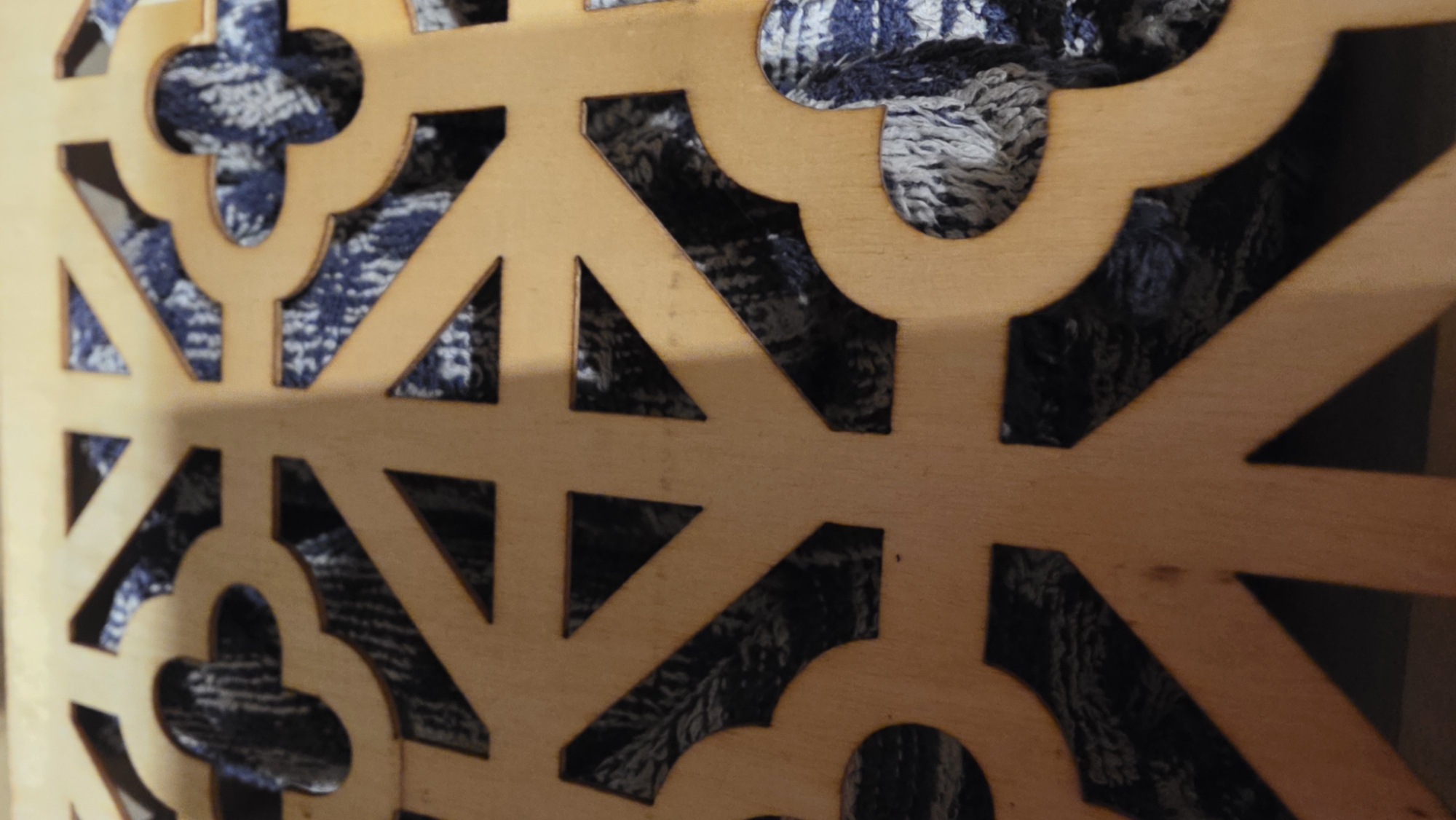 A close-up shot focuses on a light brown, geometrically patterned screen or decorative panel. The design incorporates quatrefoil and triangular shapes cut out to reveal a blue and white textured material or object behind it. The lighting is subdued, highlighting the texture of both the screen and the background object. No information is provided to conclude the location is in the city of Saint Petersburg, Russia.