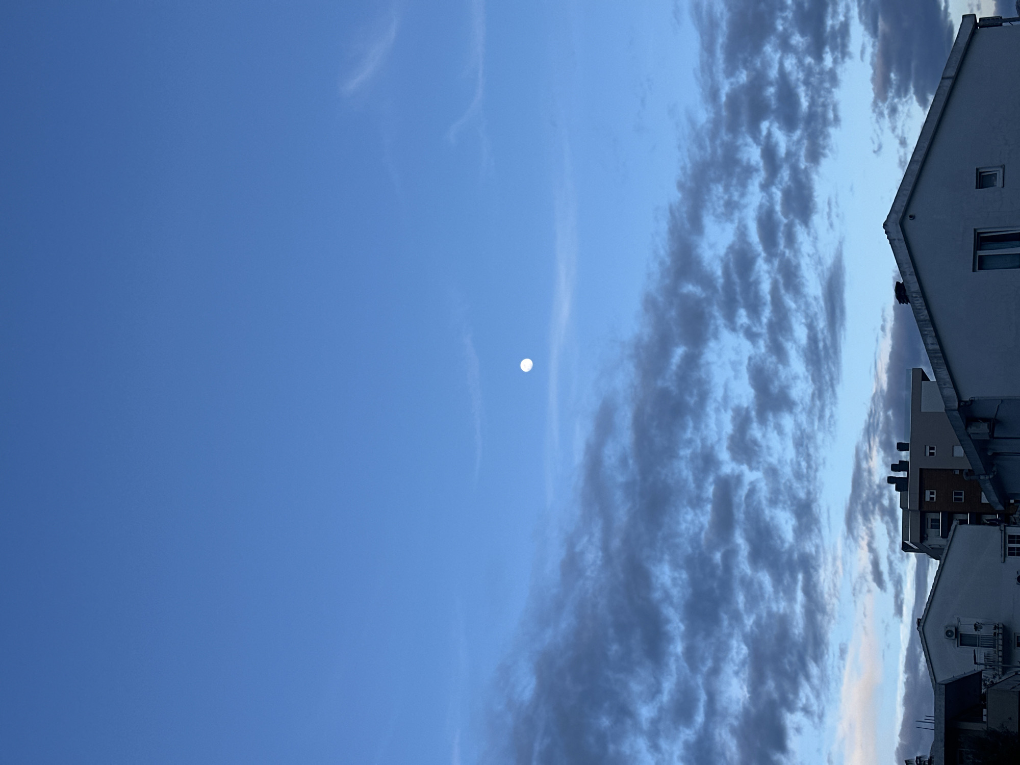 The image presents a late afternoon or early evening sky over an urban area. The left side of the image shows a clear, light blue sky with a small, bright, circular object (likely the moon or a star). To the right, the sky is filled with dense, textured clouds, ranging in color from light grey to dark blue-grey.
In the lower-right corner of the image, the tops of several buildings are visible. The buildings are white or light-colored, with visible windows and some architectural details. The image is slightly tilted, giving a vertical orientation to the scene.
Given the location information provided, it is plausible that the photograph depicts a sky view in Budva, Montenegro, though the buildings themselves do not provide conclusive evidence.