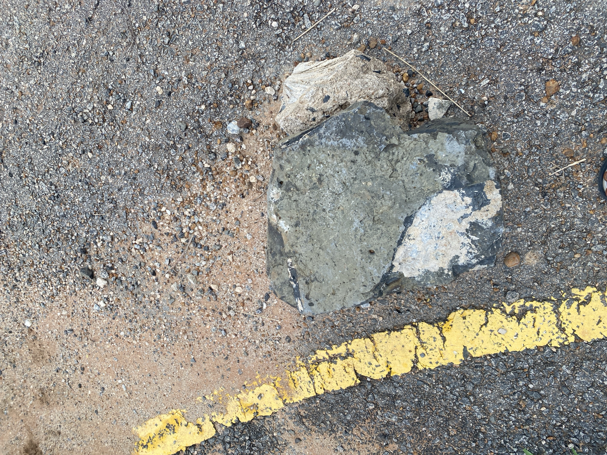 In Dumnga, Nigeria, on an unpaved surface next to a road marked with a yellow line, two large rocks lie on the ground. One rock is dark gray with lighter gray and white patches. The other rock is lighter in color, appearing to be a pale beige and white with some dark flecks. The ground is covered with a mixture of dark gravel and sand, with smaller pebbles and debris scattered throughout. A dry blade of grass lies on the ground near the rocks.