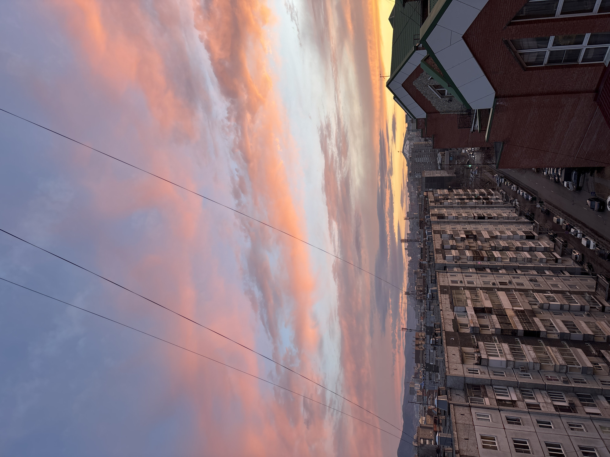 A panoramic view of an urban landscape at either sunrise or sunset, captured from a high vantage point in Krasnoyarsk, Russia. The upper two-thirds of the image are dominated by a spectacular sky, filled with long, streaky clouds displaying a vibrant palette of colors. These clouds transition from bright golden-orange and pink tones on the right, suggesting the direction of the sun, to softer purples and blues on the left. Several thin black power or communication lines traverse horizontally across the upper part of the sky.
In the lower third of the frame, an extensive city scene unfolds. On the right, a multi-story brick building with a distinctive green roof and white trim is prominent in the foreground, featuring numerous windows and architectural details. Adjacent to this building, a street is visible, lined with various parked cars. Extending into the mid-ground, a dense array of multi-story apartment blocks creates a textured urban fabric. These buildings appear to be of concrete construction, characterized by numerous windows and some visible air conditioning units. In the far distance, a range of mountains or hills is silhouetted against the colorful horizon. The scene evokes a sense of tranquility and highlights the natural beauty of a dawn or dusk sky over a bustling city. There is no visible text or specific human activity depicted.