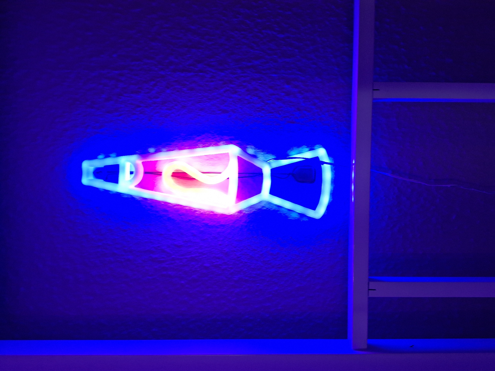 The image depicts a neon sign shaped like a lava lamp, illuminated against a textured dark blue wall. The neon tubing outlines the iconic shape of a lava lamp, with a bulbous top, a cylindrical middle, and a wider base. The colors of the neon are primarily bright pink and yellow for the "lava" within the lamp, and white and blue for the outer casing. The sign is positioned centrally in the frame, casting a vibrant glow on the surrounding area. A white shelf or ledge is visible in the foreground, below the neon sign. A thin black wire, likely part of the sign's power supply, dangles down from the base of the lava lamp neon and hangs near the shelf. The overall ambiance is dim and atmospheric, dominated by the intense colors of the neon light. The image does not contain any information that would allow for its location to be identified in the city of Wildeshausen, Germany.