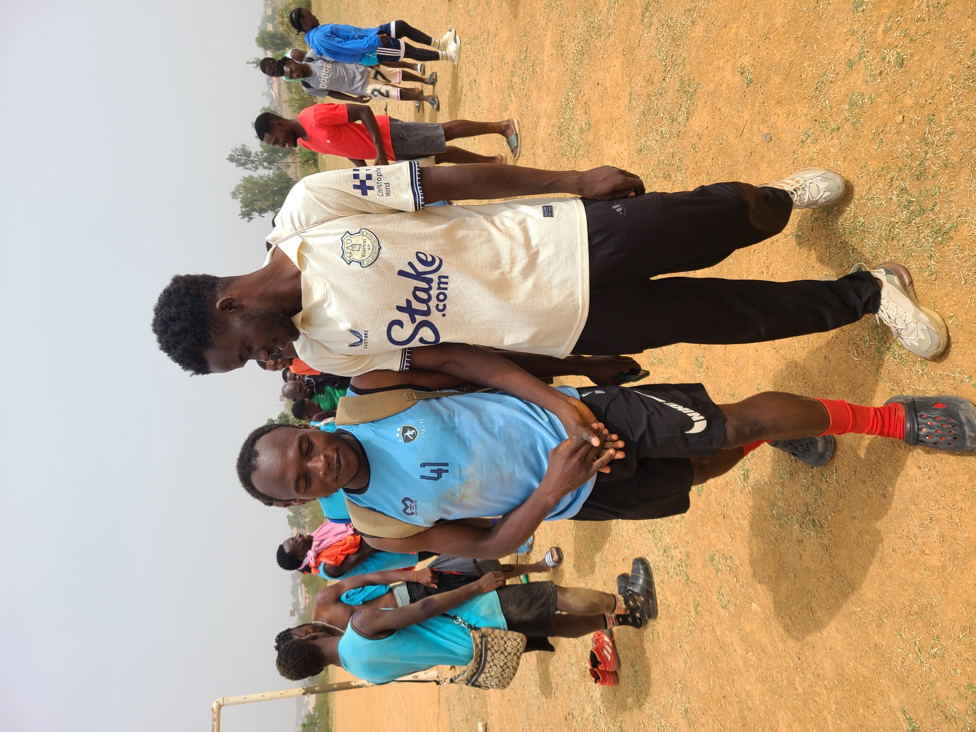 The image shows a group of young men on a dirt field in Barikin Jirga Summa, Nigeria. Two men are in the foreground. One is wearing a light blue soccer jersey with the number 41, black shorts, red socks, and black clogs. The other is wearing a white and gold soccer jersey with "Stake.com" printed on it, black athletic pants, and white sneakers. They are standing close to each other. In the background, several other men are visible wearing various colored shirts and shorts. A goalpost is partially visible at the edge of the frame. The sky is overcast, and the ground is dry and dusty.