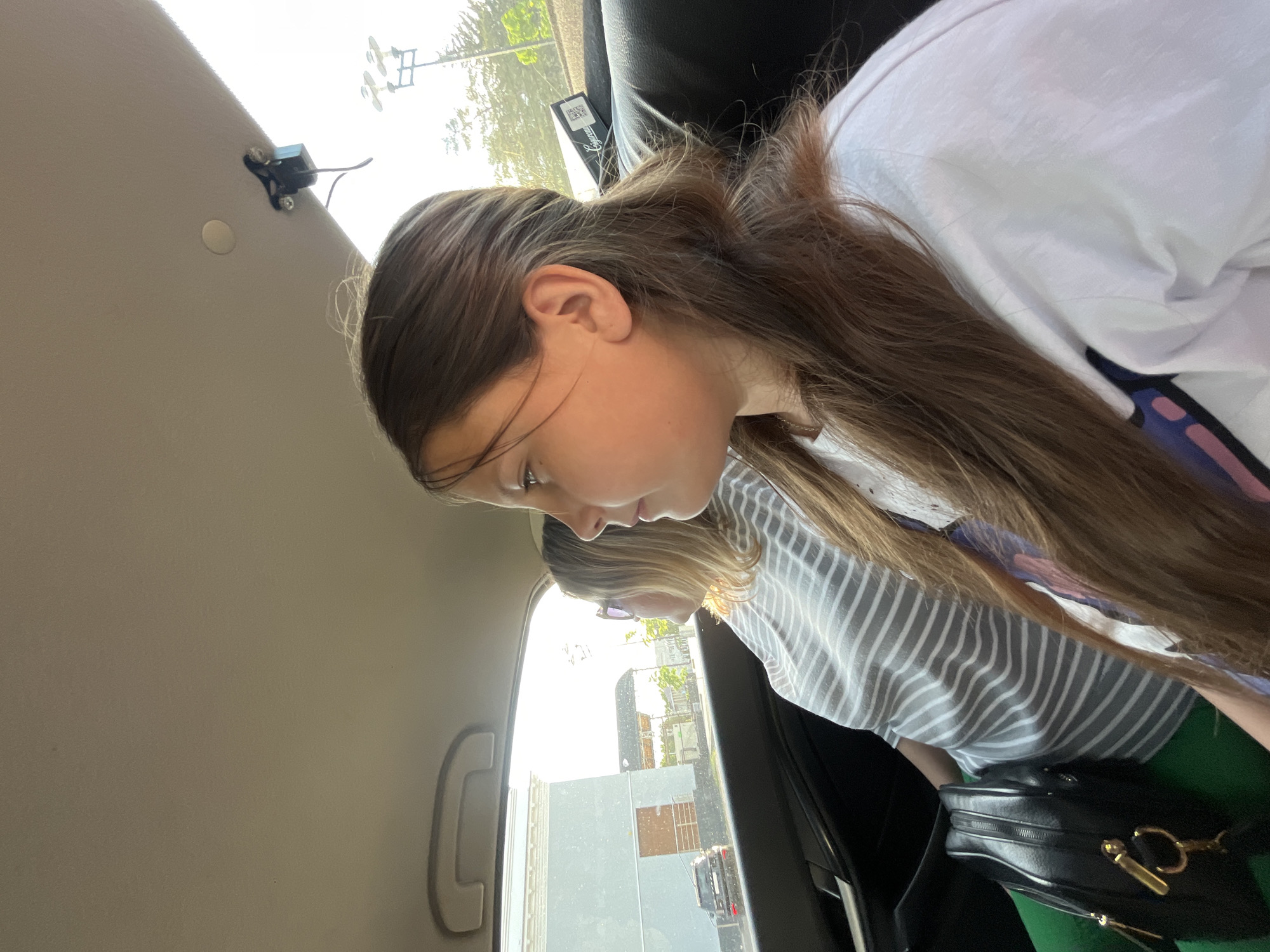A view inside a car shows a young girl with brown hair, wearing a white shirt with a blue and pink design, sitting in the back seat. Another individual with blonde hair wearing a striped shirt is also present, partially visible behind the girl. A black purse is near them. Through the car window, buildings and streetlights are visible, suggesting an urban area, though I cannot confirm the specific location of Novopokrovskaya, Russia without more information. The interior of the car includes a handle, a ceiling light fixture, and a black headrest.