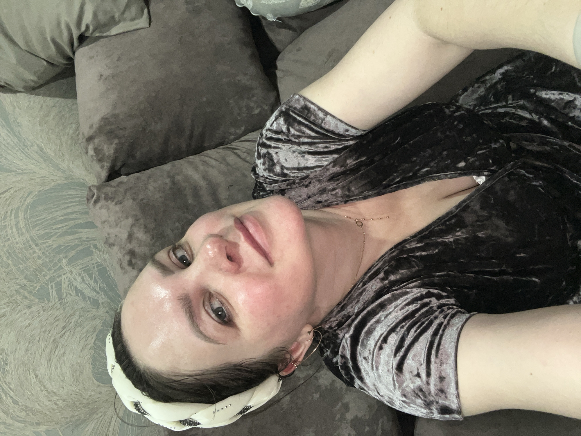 A woman is lying on a grey velvet sofa in what appears to be an indoor setting. She is wearing a dark velvet dress and a white patterned headscarf. The wallpaper is light-colored with a faint design. A gold necklace and multiple earrings are visible. The woman's face appears flushed, and her eyes are open. No details suggest this is in the city of Noviy, Russia.