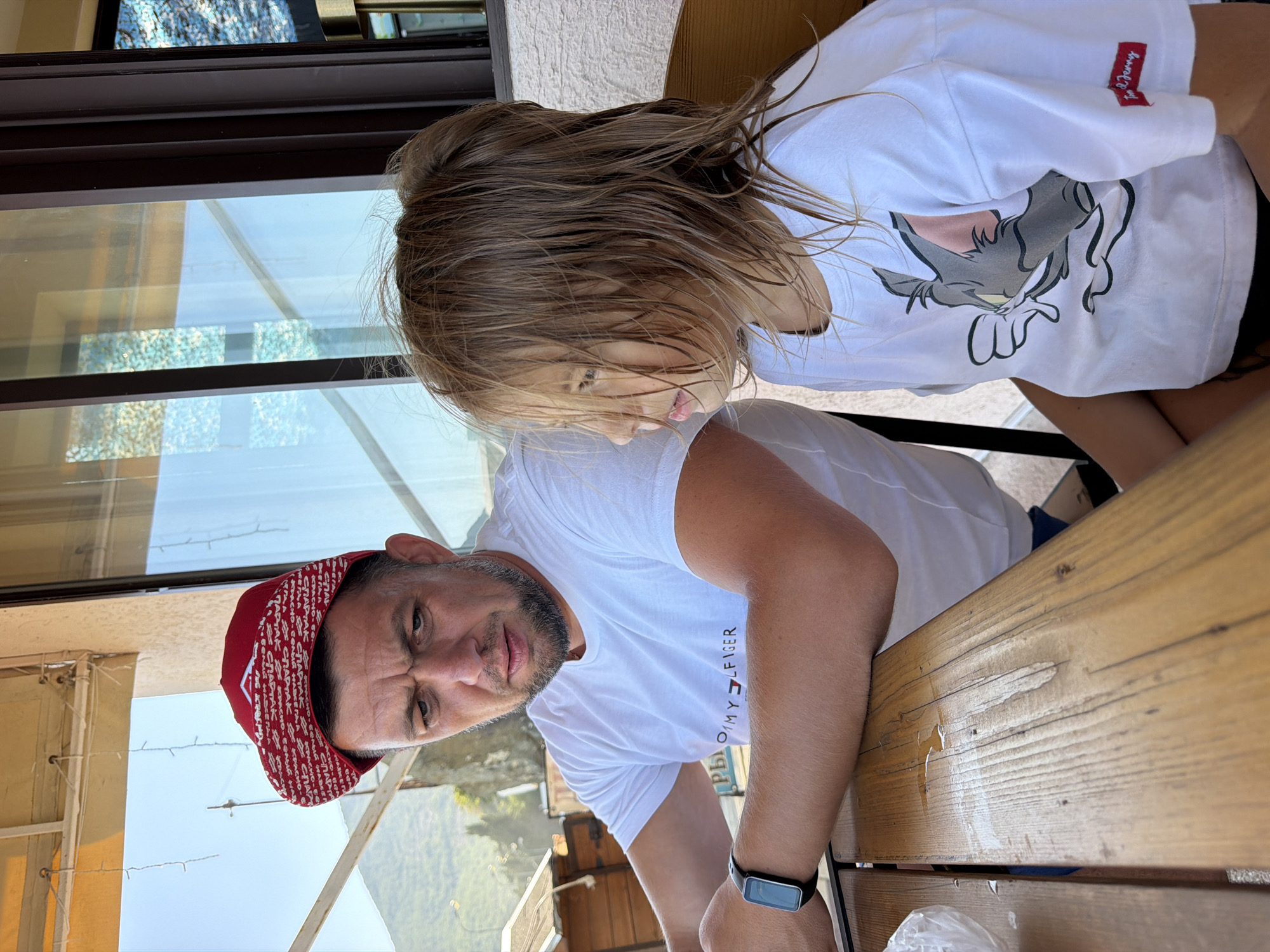 A man wearing a red cap and a white t-shirt with "Tommy Hilfiger" branding is seated at a wooden table. He has a beard and appears to be looking downwards. A young girl, also wearing a white t-shirt with a Tom and Jerry graphic, is seated to his right, looking away from the camera. They are outdoors, likely on a terrace, with a window and foliage visible in the background. The environment suggests a casual setting, possibly a cafe or restaurant. The overall scene captures a moment of interaction or shared presence between the two individuals.