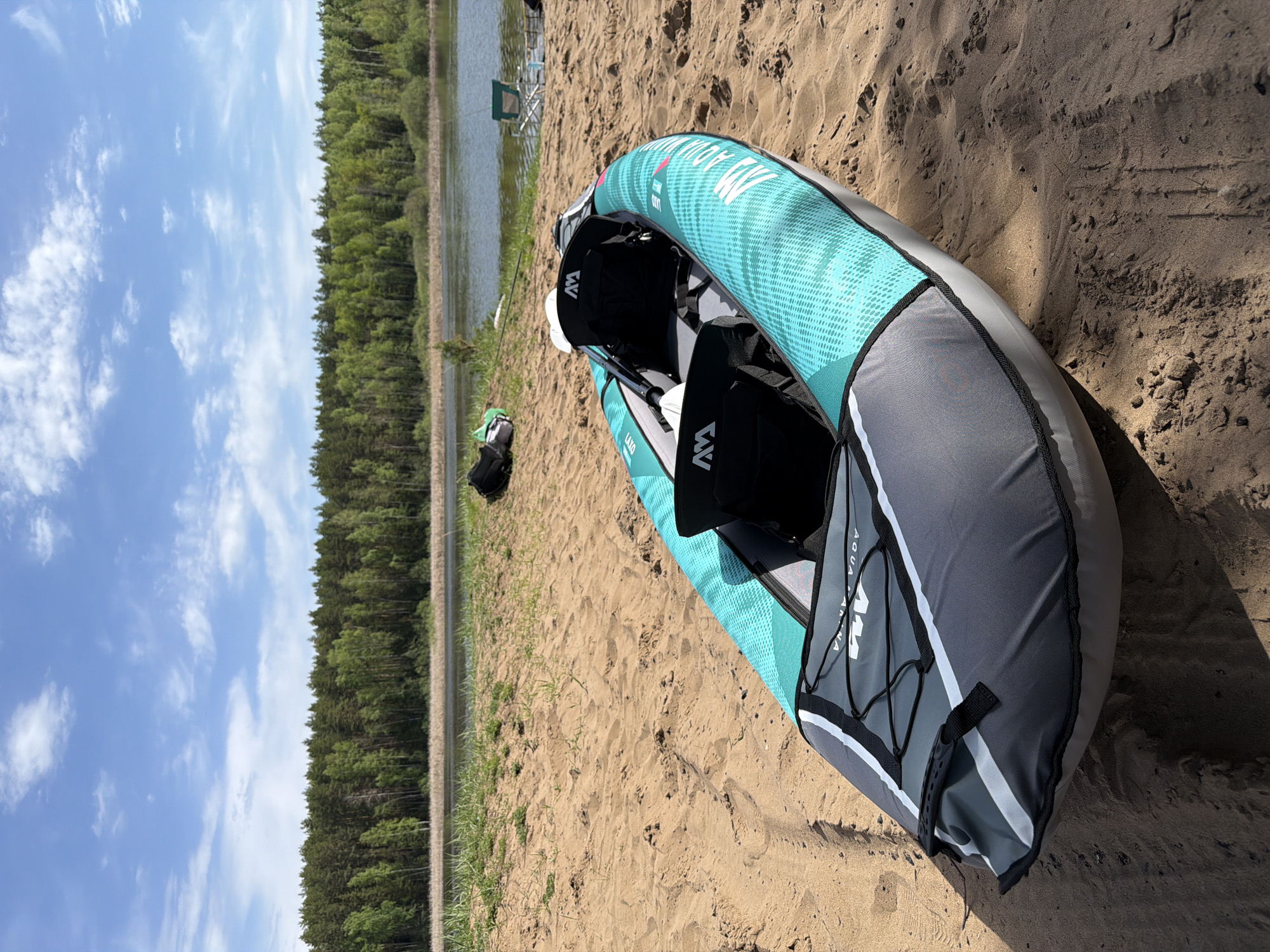 The image depicts an inflated Aqua Marina LAXO-320 kayak resting on a sandy beach. The kayak is predominantly turquoise and gray, with black seating visible. The beach appears to be located on the edge of a lake or river, with a line of trees in the background and a bright sky overhead. In the distance, a fishing chair and pole can be seen. There are also some bags close to the kayak. Based on the environment shown in the image, it is possible this photo was taken in the city of Chaikovsky, Russia.